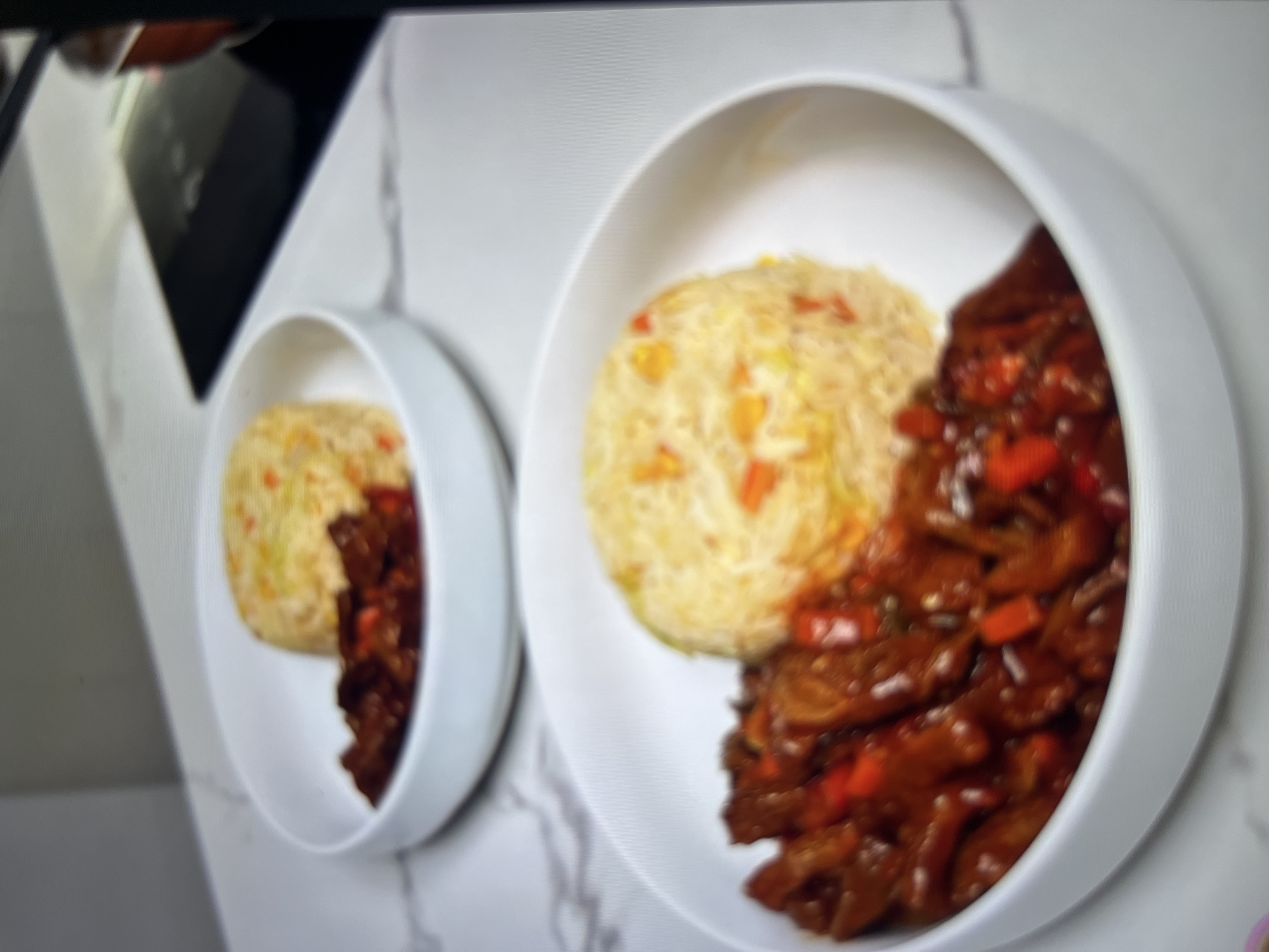 The image shows two white bowls of food on a white marbled countertop. Each bowl contains fried rice with visible pieces of carrots and peas, and a side of what appears to be beef in a dark sauce with red bell peppers. The food is arranged to create a neat presentation. In the background, a black induction cooktop is partially visible. The scene is clean and well-lit, suggesting a domestic kitchen environment. There are no people or animals present in the image. The overall impression is of a prepared meal ready to be served. The image does not provide any specific details or clues that would allow for the localization of the scene in Abuja, Nigeria.