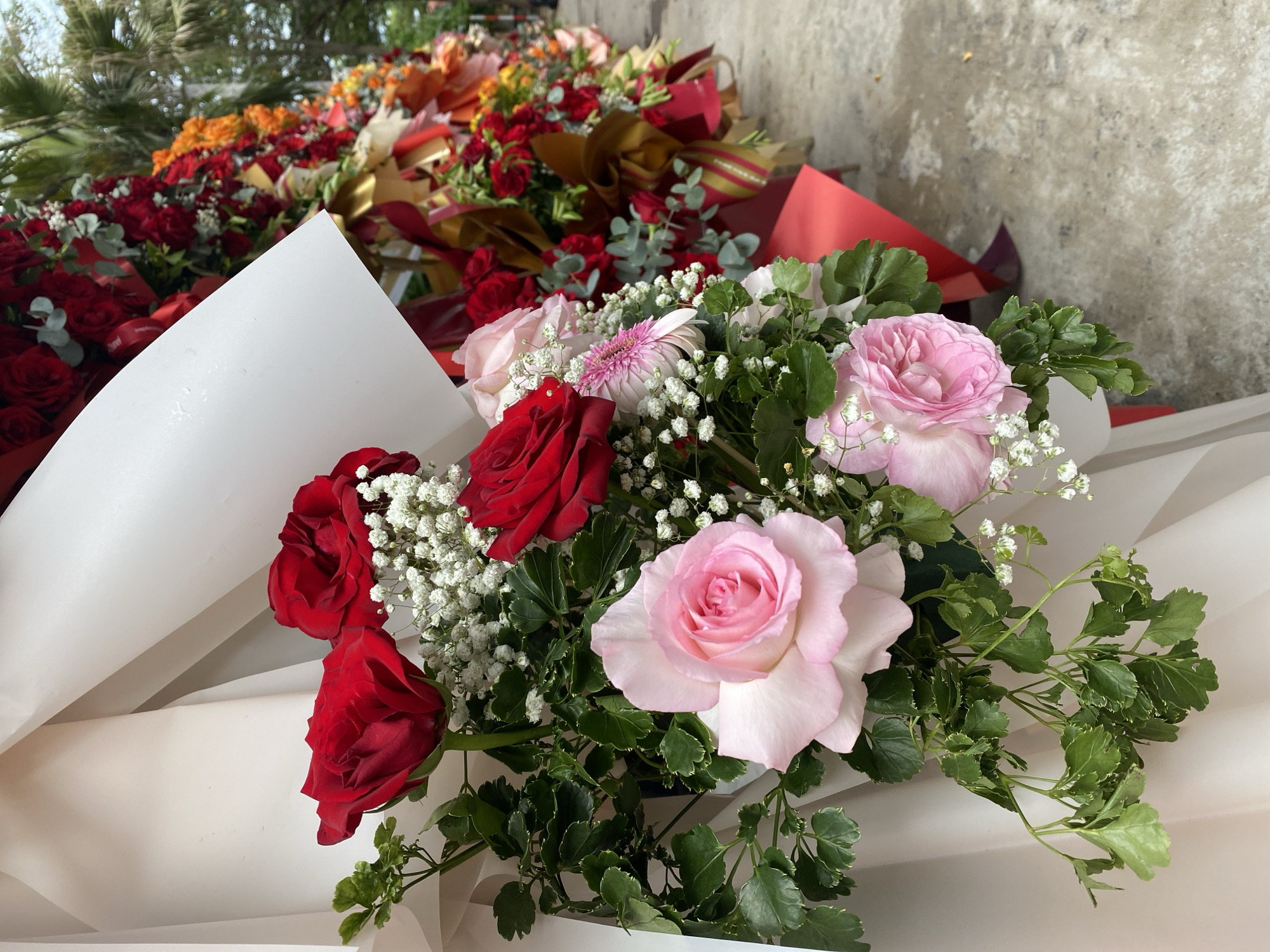 The image depicts a close-up view of a vibrant floral arrangement, predominantly featuring red and pink roses, interspersed with delicate white baby's breath and lush green foliage. The flowers are presented in a decorative manner, likely for a special occasion or as part of a larger display.
In the background, a line of similar floral arrangements extends into the distance, suggesting an outdoor event or public space decorated with flowers. The background also shows hints of palm trees and a cloudy sky, indicating an outdoor setting. The ground appears to be paved, possibly a sidewalk or pathway.
Based on the context of the image and common knowledge of flower displays in Đà Nẵng, Vietnam, this scene is likely located along a street or public square during a festive period, such as Tết (Vietnamese Lunar New Year) or a wedding. Such elaborate floral decorations are often seen adorning businesses, public buildings, or along main thoroughfares in Đà Nẵng to create a celebratory atmosphere. The specific location within the city is not identifiable from the image alone.