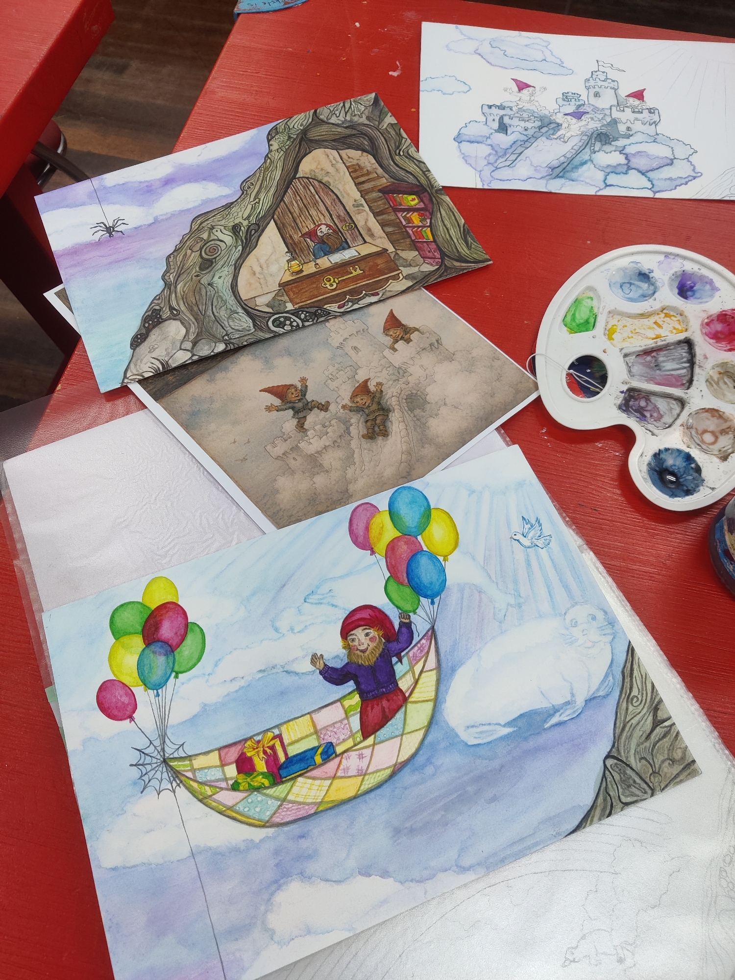 This media file displays several watercolor paintings laid out on a red surface. The paintings feature whimsical, gnome-like characters and fantasy scenes.
One painting depicts a gnome with a red hat and beard, wearing a purple tunic, floating in a hammock-like basket suspended by balloons. The hammock contains wrapped gifts. Beside the hammock, a white seal-like creature is visible, and a small white bird is drawn in the sky.
Another painting shows a gnome at a desk inside a hollowed-out tree trunk, with bookshelves in the background. A spider is shown suspended from a thread outside the tree.
A third painting illustrates a castle perched on clouds, with three gnomes in red hats in front of it. A bridge connects the castle to the clouds.
A watercolor palette with various colors, including green, blue, purple, and pink, is placed on the red surface next to the paintings. The scene appears to be an art workspace, possibly in Winterswijk, Netherlands. The overall mood of the artwork is lighthearted and fantastical. No visible text is present in the media file.