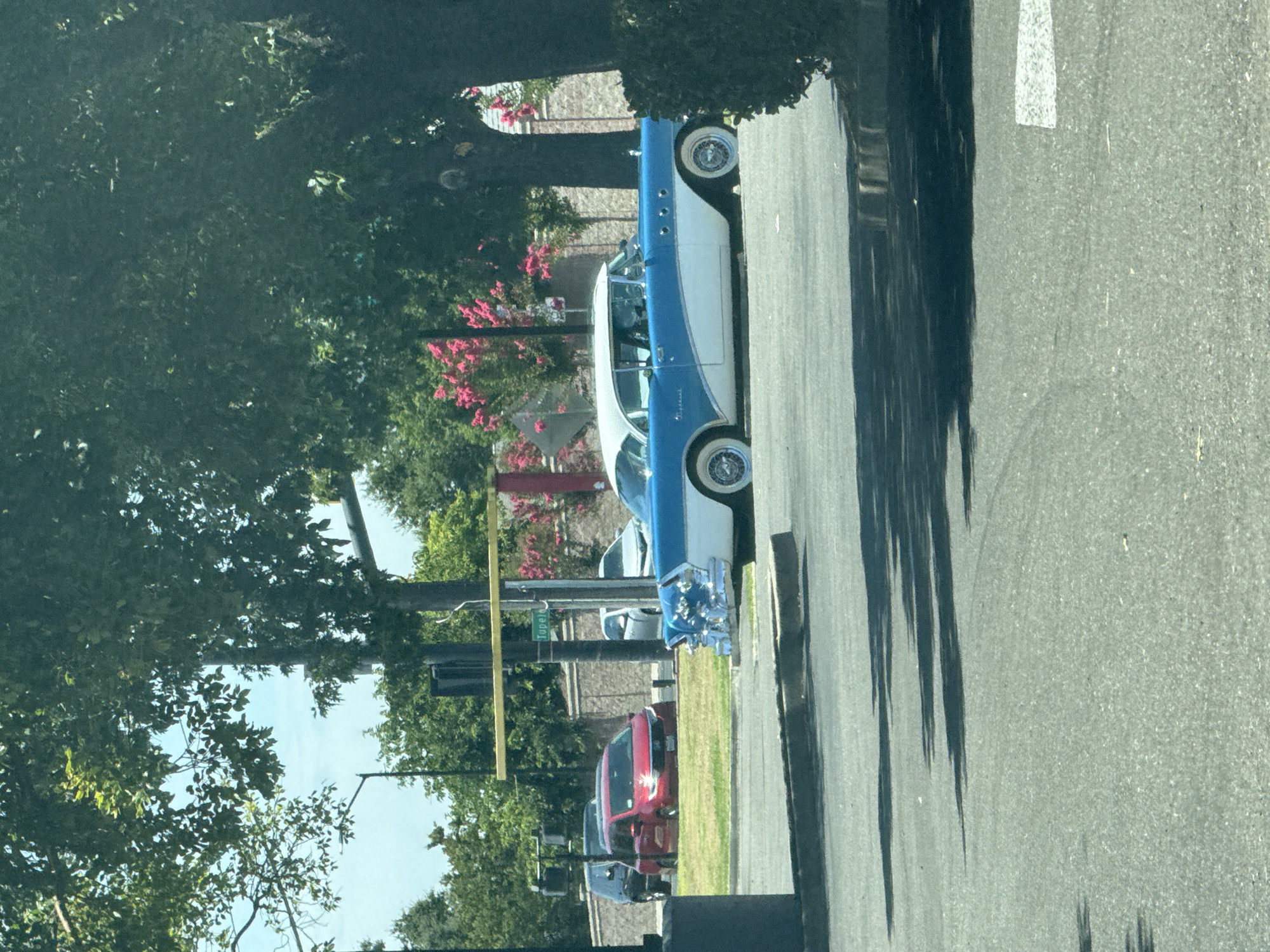 In Citrus Heights, United States of America, a vintage two-tone blue and white Buick sedan is parked on asphalt. The car is positioned in the foreground, angled slightly to the right, with its chrome detailing prominently visible. Behind the Buick, a red minivan is parked on a grassy area, and further back, a dark SUV is also visible. A street sign that reads "Tupelo" hangs on a yellow support structure. The scene is framed by lush green trees, with some pink flowering bushes in the background. The sky is bright and appears to be daytime. Shadows from the trees are cast across the asphalt.