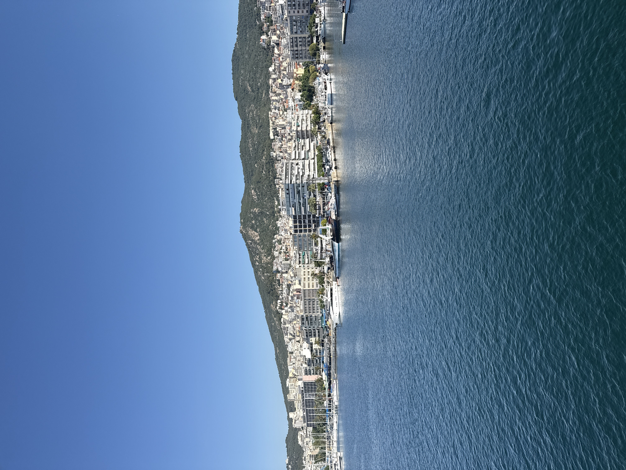 This image shows a view of the harbor and waterfront of Kavala, Greece. In the foreground, the blue water of the harbor is calm with small ripples. Moored boats of various sizes, including yachts and sailboats, are visible along the waterfront. Behind the boats, a strip of land features buildings, ranging from modern structures with white facades to older, more traditional architecture. These buildings climb up the side of a hill that forms the backdrop of the city. The hill is covered in greenery and also dotted with buildings. The sky above is a clear, bright blue, indicating a sunny day. The scene is one of a bustling port city situated against a scenic natural landscape.