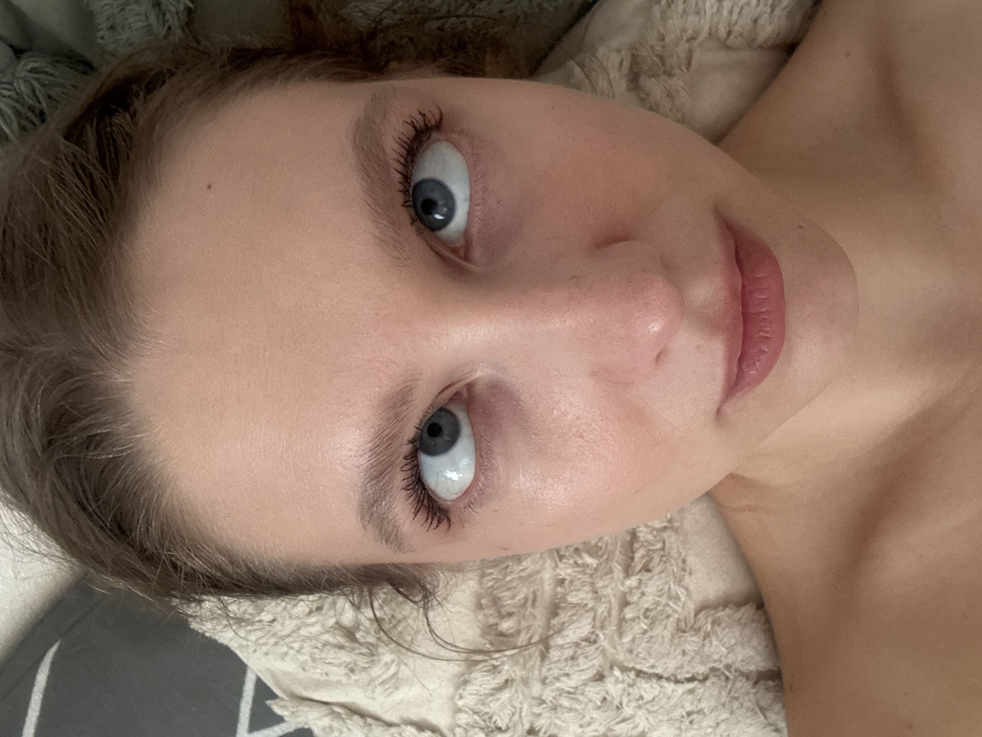 The image is a close-up, high-angle shot of a young woman's face, looking upwards. Her eyes are crossed, and she is wearing mascara. Her lips are slightly parted, and she has a mole on her forehead. She appears to be lying down on a textured surface, possibly a blanket or pillow, with hints of a grey patterned fabric in the background. The lighting is soft, illuminating her skin and features. There are no visible indicators in the image that would allow for the determination of its location within the city of Izhevsk, Russia.