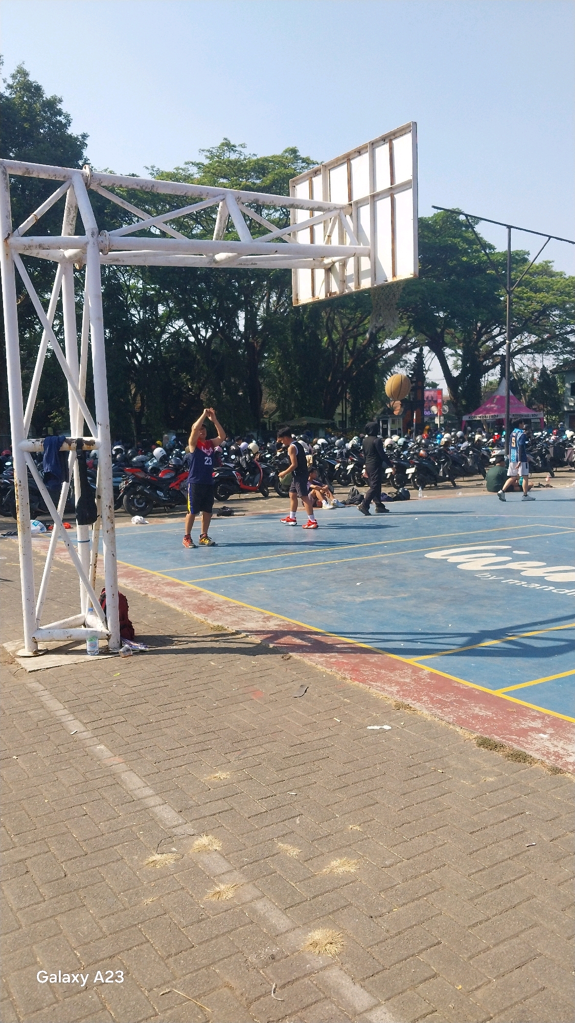 The image depicts an outdoor basketball court, likely located in the city of Malang, Indonesia, under a clear blue sky. The court has a blue surface with red and yellow boundary lines. In the foreground, a white metal basketball hoop structure stands on the left side of the frame. Several people are visible on the court, engaged in or observing basketball-related activities. One individual in the mid-ground, wearing a blue tank top with the number "23" and black shorts, is standing with arms raised. Another person in a black tank top and shorts is also on the court. In the background, a multitude of parked motorcycles are arranged, suggesting a public space with amenities for visitors. Trees line the perimeter of the court, providing shade. The overall scene is one of recreational activity in a public park or sports complex.