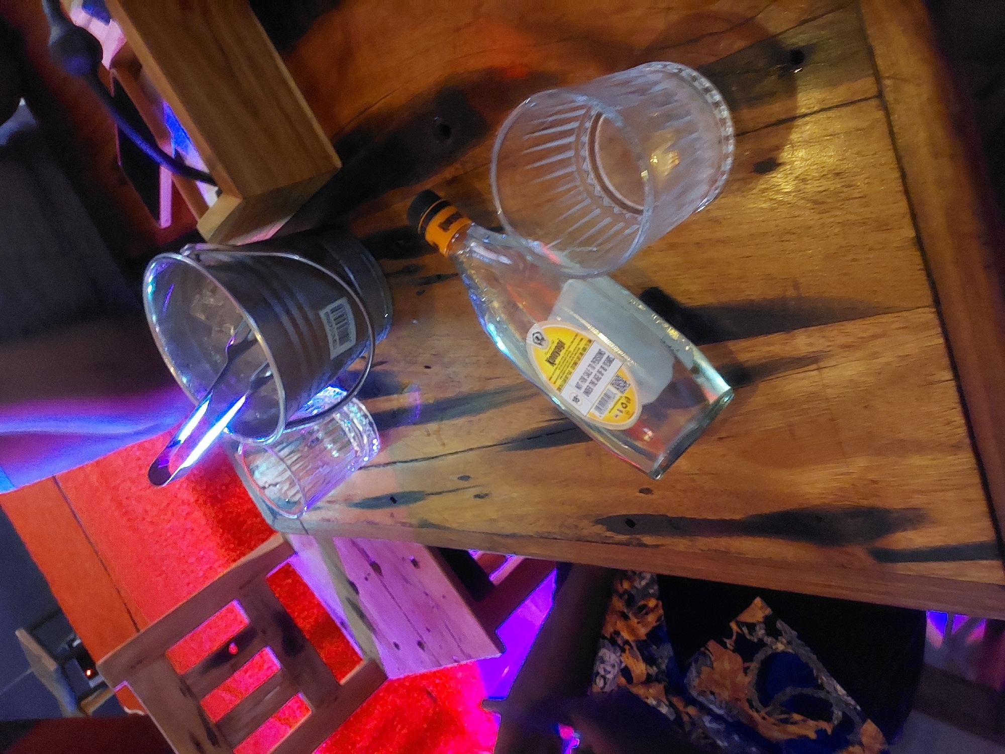 The image shows a tabletop scene, possibly within an establishment in Dar es-Salaam, Tanzania. The table is wooden and weathered with some dark markings. Present on the table are a metal bucket, a glass with remnants of liquid, an empty clear glass, and an unlabeled bottle with an orange cap. The bucket contains an elongated silver object. Below the table, there's a glimpse of a chair with a colorful pattern and what appears to be colored lighting (pink, red, and blue). A portion of a wooden frame is seen on the left side of the image.