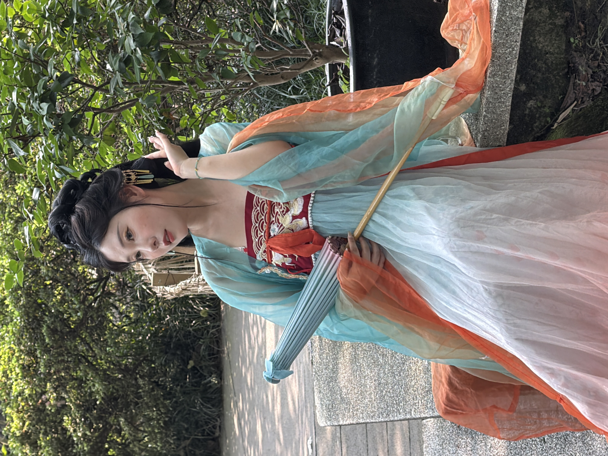 A young East Asian woman is seated outdoors in Yuzhong District, China, on a light grey, textured concrete bench or wall. She is positioned slightly to the right of the frame's center, facing left with her gaze directed towards her left. Her dark hair is styled elaborately into a high bun on top of her head, adorned with gold-toned, dangling ear ornaments featuring light blue accents. Her right hand is raised near her face, with a thin, beaded teal bracelet visible on her wrist. She is wearing a traditional Chinese Hanfu-style outfit consisting of multiple layers. An inner top is visible in red with intricate gold embroidery and a white pearl-like trim along the neckline, secured with an orange sash. Over this, she wears flowing, sheer fabric in gradient shades of teal/aqua and orange/peach, draped over her shoulders and arms. Her skirt is a light blue and white gradient, also made of flowing, sheer fabric. In her left hand, she holds a closed, light blue parasol with a wooden handle, angled across her lap. The background features lush, dense green foliage, comprising trees and bushes, suggesting a garden or park setting. To the left of the bench, a light-colored paved ground is visible. To the right and below the bench, darker ground with sparse foliage and exposed soil is present. The scene is illuminated by natural daylight.
