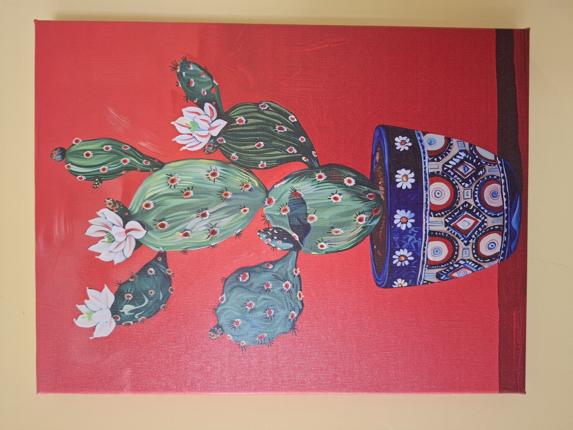 A painting of a prickly pear cactus in a decorative pot is displayed against a bright red background. The cactus has several pads, each adorned with small red and white flowers. The pot itself is a vibrant blue, featuring intricate patterns of circles and geometric shapes in various colors. The painting appears to be on canvas and is framed by a dark red border at the bottom. The artwork is hanging on a light beige wall. There are no people or other objects in the scene. This image is not located in Nha Trang, Vietnam.