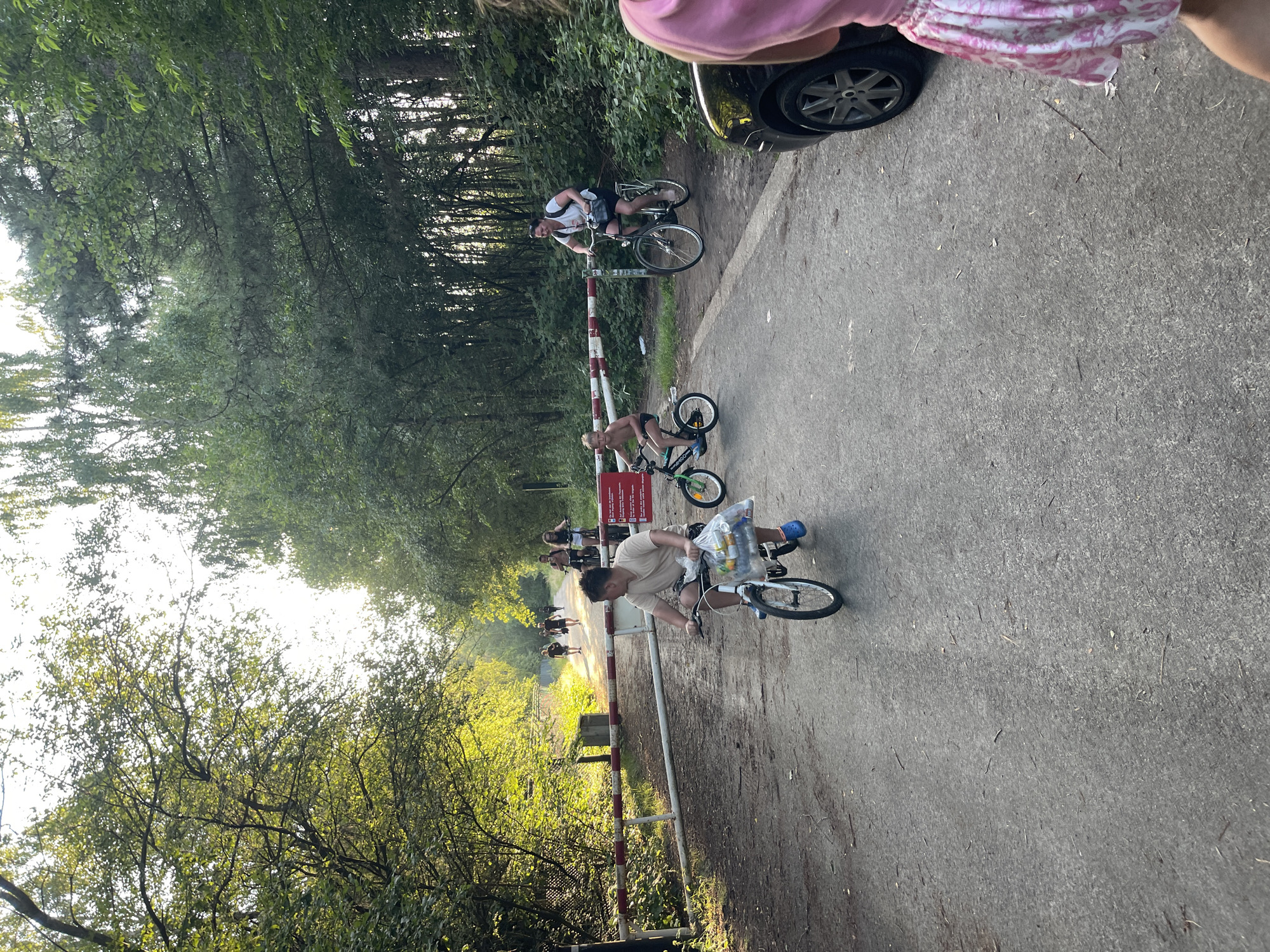 The image depicts a paved path in a wooded area, possibly a park or nature reserve, with a barrier across it. Several people are present, some on bicycles. In the foreground, a young person on a bicycle is riding towards the camera, carrying a plastic bag filled with items. To their right, another child on a smaller bicycle is also riding. Further down the path, a woman on a bicycle is visible, and in the background, several other individuals are walking or cycling. A black car is parked on the right side of the path. The trees on either side of the path create a canopy overhead, with sunlight filtering through the leaves. The overall scene suggests a recreational outing in a natural setting.