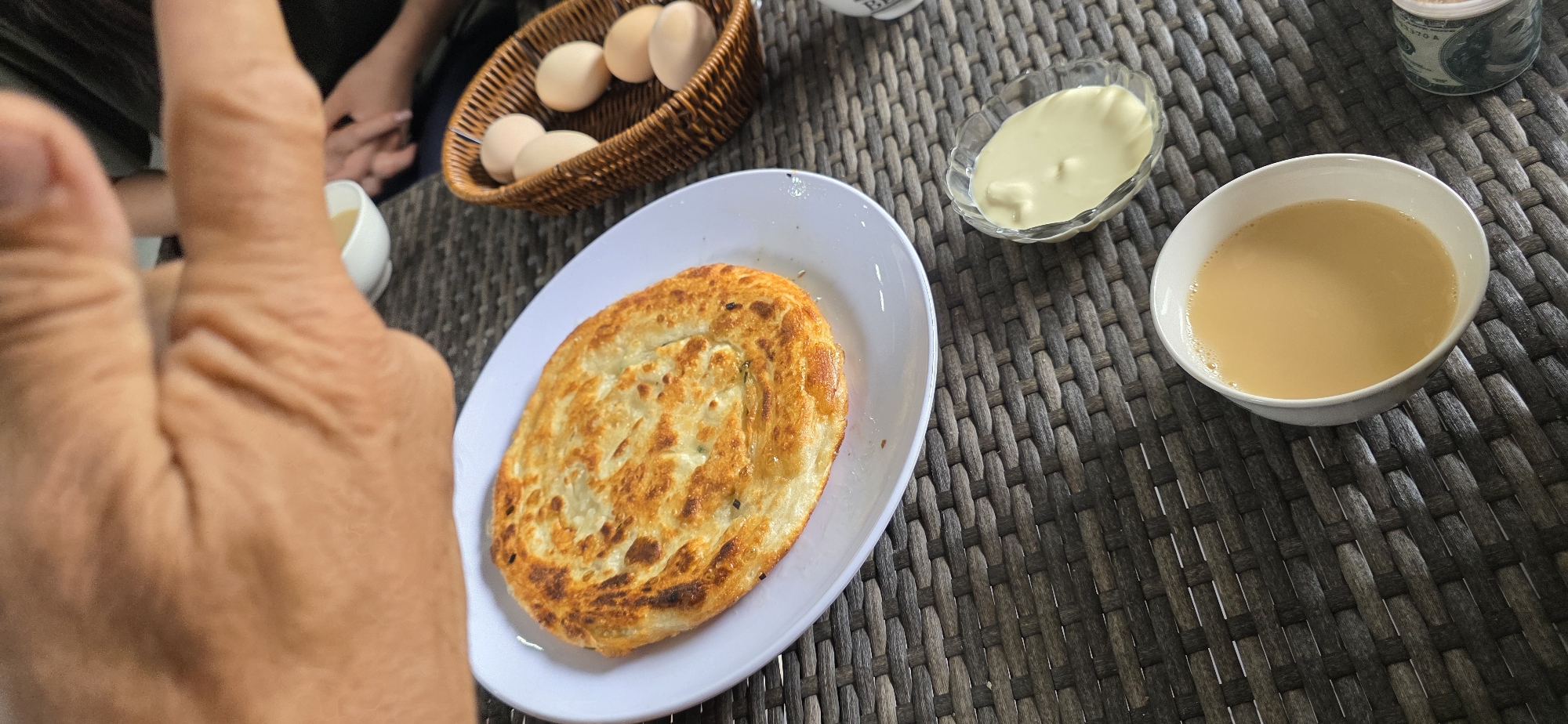 A close-up, angled shot shows a table setting with food and beverages. In the foreground, a white plate holds a golden-brown, layered flatbread. To the right of the flatbread, a small glass bowl contains a pale yellow, creamy substance. Further to the right, a woven basket holds several light brown eggs. A white ceramic mug filled with a light brown liquid, possibly tea, is positioned in the lower right quadrant of the image. The table surface is a dark woven material. In the upper portion of the image, a person's hand, with visible wrinkles, is partially visible, suggesting someone is reaching for or interacting with the food. In the background, out of focus, another person's hands and arms can be seen. The environment appears to be outdoors, possibly a cafe or restaurant in Sasyk-Bulak, Kyrgyzstan.