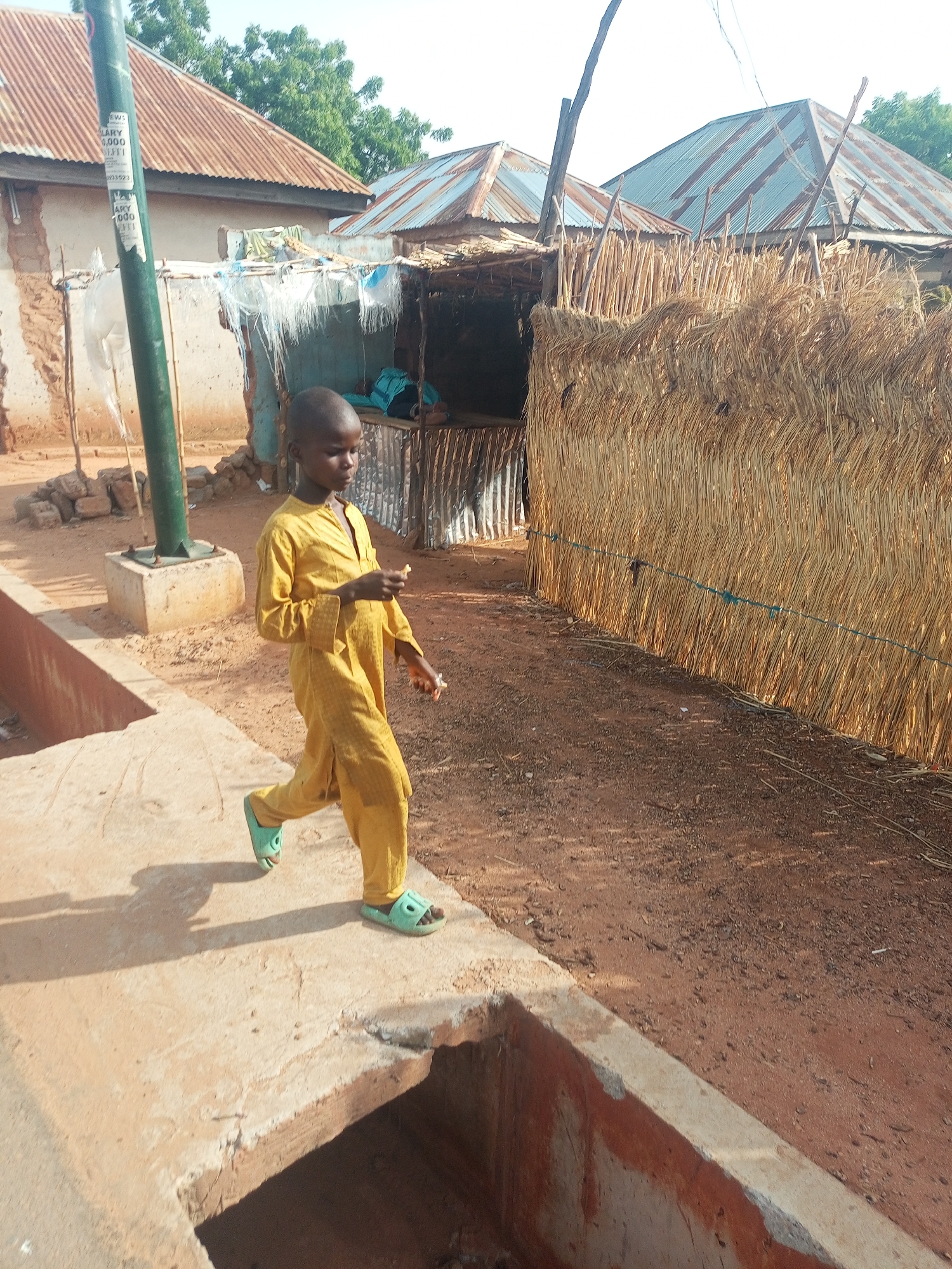 A child in a yellow jumpsuit and green sandals walks on a concrete curb near a drainage ditch in Gombe, Nigeria. The child is holding something in their hands. In the background, there are buildings with corrugated metal roofs and a hut constructed from sticks and hay. A green pole is visible near one of the buildings. The ground is dirt.