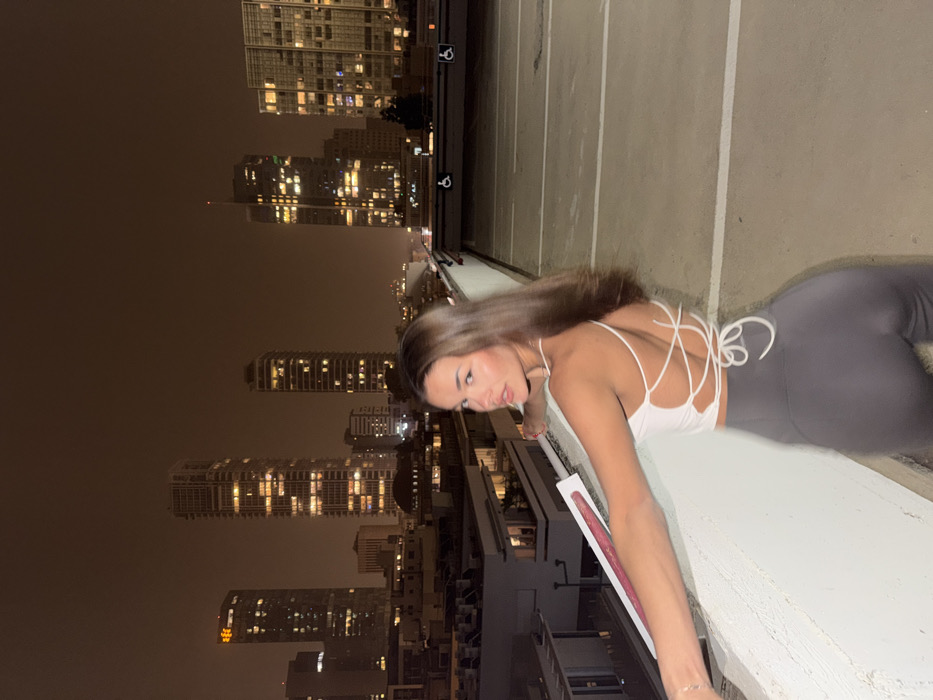 The image captures a single young adult female posing at night on what appears to be the rooftop or upper deck of a multi-story parking structure in Tel Aviv, Israel. She is positioned with her back partially towards the camera, looking over her left shoulder directly at the viewer. Her left arm is extended, resting on a light grey concrete barrier that demarcates the edge of the elevated platform. She holds a slender white object with a red-pink stripe on its side in her left hand. The female subject wears a white, backless top with multiple criss-crossing straps tied at the lower back, paired with dark grey form-fitting pants. Her hair is dark brown and styled loosely around her shoulders. The background displays a densely built urban environment at night, under a dark, light-polluted sky. Numerous illuminated high-rise buildings are visible, including one prominent skyscraper in the center, identifiable as the Azrieli Sarona Tower, consistent with the Tel Aviv skyline. The ground surface of the platform is light grey concrete, showing faint white markings indicative of parking spaces. The subject is brightly illuminated, suggesting a direct light source, contrasting with the darker city lights behind her.