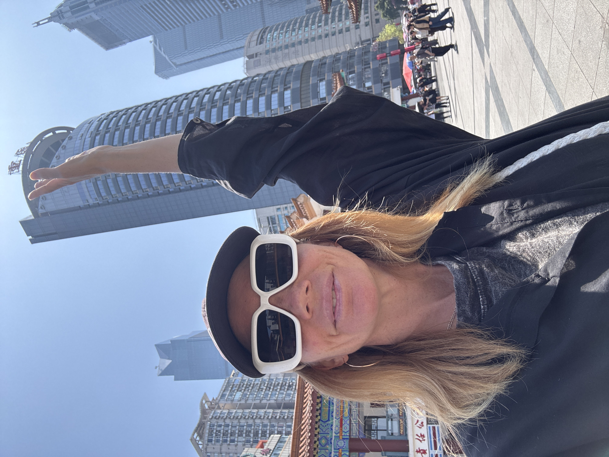 A woman is photographed at eye-level from a close-up perspective, appearing to gesture towards tall buildings in the background. The woman is wearing a dark-colored brimmed hat and white-framed sunglasses, with blond hair falling around her face. She has a fair complexion and is wearing a dark-colored shirt or jacket.
In the background, there are multiple modern skyscrapers. Some of the buildings have a cylindrical design, while others are more angular. At the base of the photograph, there is the edge of a traditional Chinese building with vibrant colors and intricate details.
The scene is set in Yuzhong District, China, evidenced by the combination of modern architecture and traditional Chinese elements. A crowd of people is visible further into the background, suggesting a public space or square. The sky is a clear blue, indicating a sunny day.