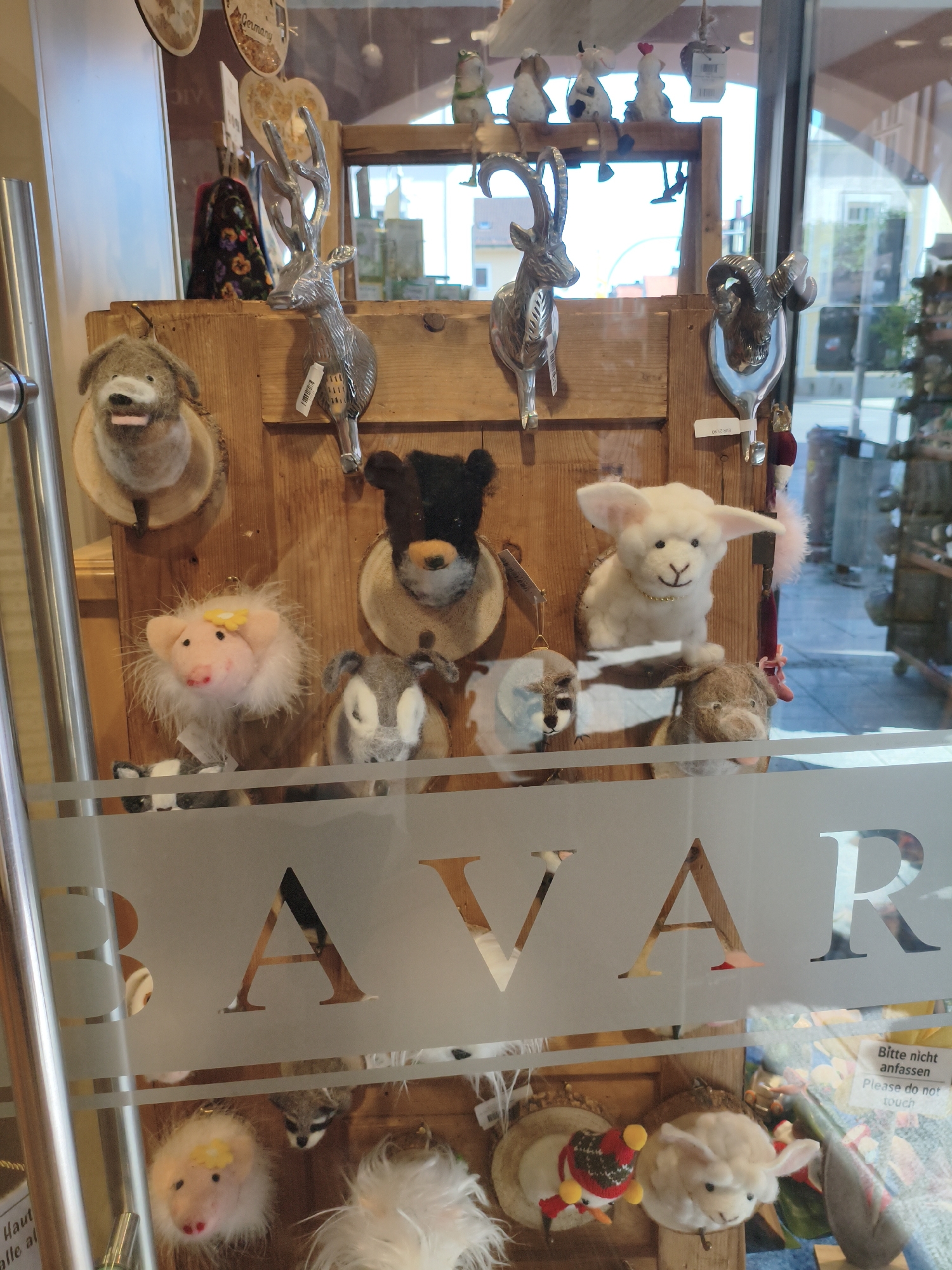 The image shows a display of novelty animal figures in a shop window in Füssen, Germany. The display features several felt animal heads, including a dog, a pig, a sheep, and a cow, as well as metal deer and ram head wall mounts. Behind the animal figures, a wooden shelf unit is visible, with decorative items and other merchandise on display. A large "BAVAR" logo is etched into the glass of the shop window, suggesting the shop is located in Bavaria. A sign with German and English text reads "Bitte nicht anfassen. Please do not touch."