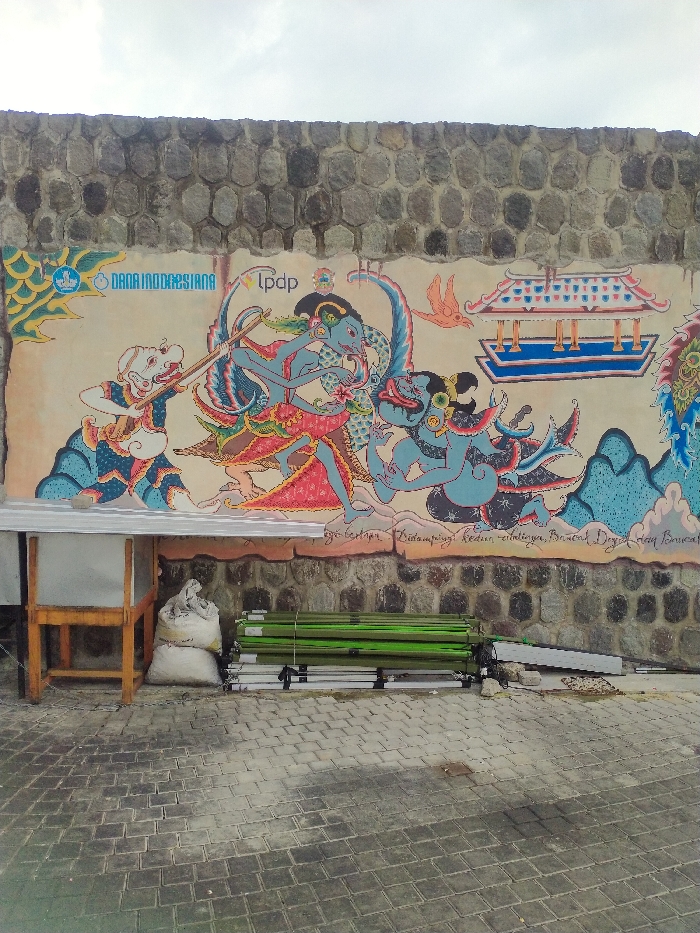 An outdoor scene features a vibrant mural painted on a stone wall under an overcast sky. The wall itself is constructed from large, irregularly shaped grey stones.
The mural, rendered in a style reminiscent of traditional Indonesian Wayang art, depicts several figures in dynamic poses against a light background. On the far left, a light-skinned character, possibly a Punakawan (clown figure from Javanese shadow puppet theater), is shown holding a long staff or spear. To its right, a central blue-skinned figure, likely a deity or hero, wears an elaborate headdress and garments, and appears to be drawing a bow, with a green-feathered wing-like element behind its head. Further right, another blue-skinned character, possibly a demon or another powerful figure, is also depicted in an active pose. A traditional Indonesian pavilion-style building with a red roof and blue base, an orange bird, and stylized blue mountain shapes complete the background elements of the mural.
Visible text on the mural includes:
* On the upper left: A blue and white logo (featuring a stylized figure or boat) next to the words "DANA INDONESIANA".
* On the upper center: The letters "lpdp" in lowercase.
* Along the bottom of the mural, a line of handwritten text in Indonesian is partially legible. Parts read "yang bertau.", followed by "Didampingi kedamaian," and then less clear words, which the OCR suggests include "kedun dingen, Binnend Dryill day lonuca."
In the foreground, to the left, there is a simple wooden stand or table with a white surface, possibly covered by a tarp. Beneath it, two white sacks are stacked. To the right, a pile of folded green items, resembling chairs or small tents, are stacked horizontally on the ground, alongside some silver-colored metal frames. The ground is paved with grey rectangular interlocking blocks, indicating a public or semi-public outdoor area.