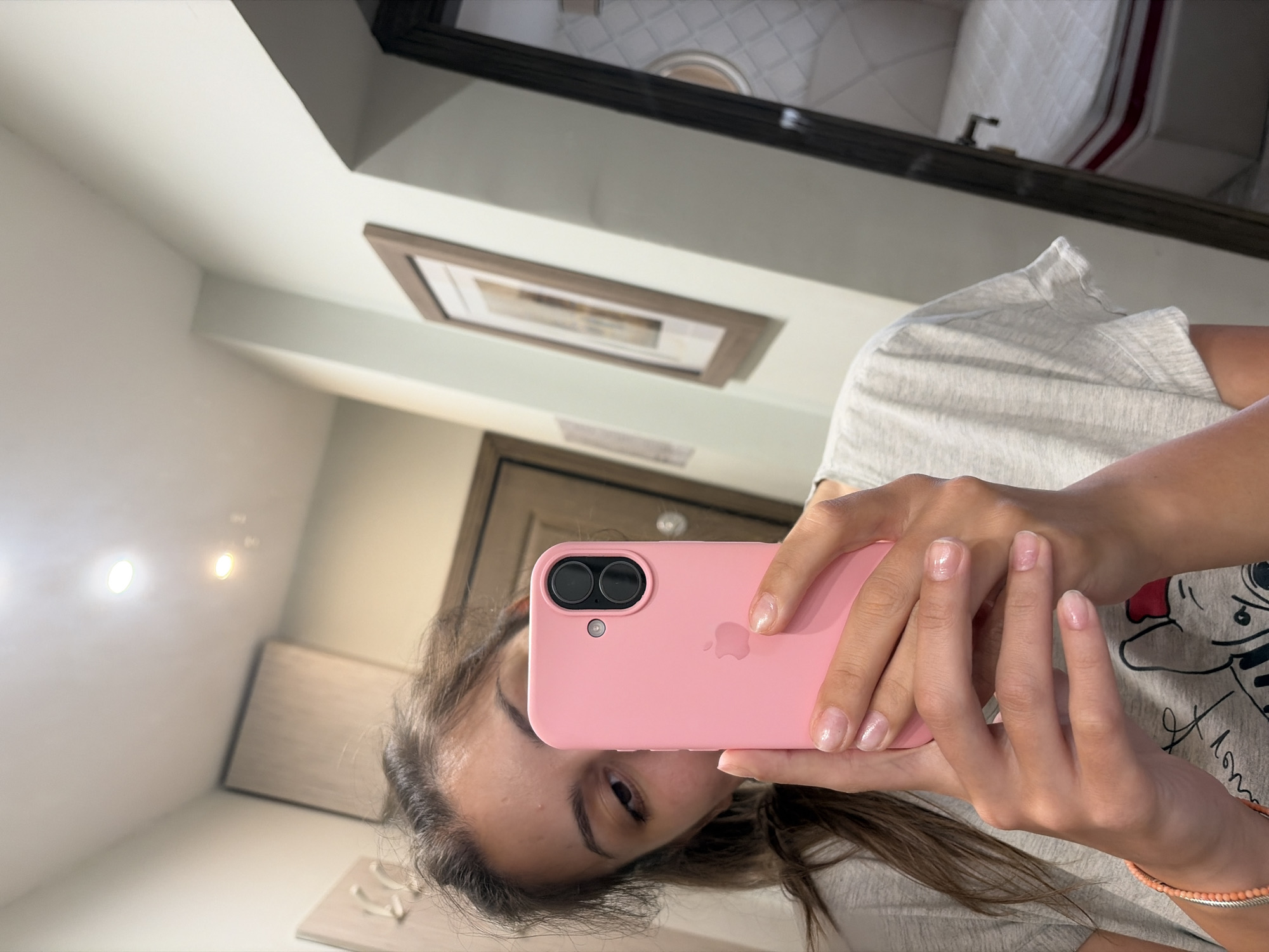 A young woman is taking a selfie in a bathroom. She is holding a pink iPhone in her left hand, which is obscuring her face. She is wearing a grey t-shirt. The bathroom has white walls and beige cabinets. There is a framed picture on the wall and a mirror reflecting a bed. The image is taken in a bathroom in Burgas, Bulgaria.