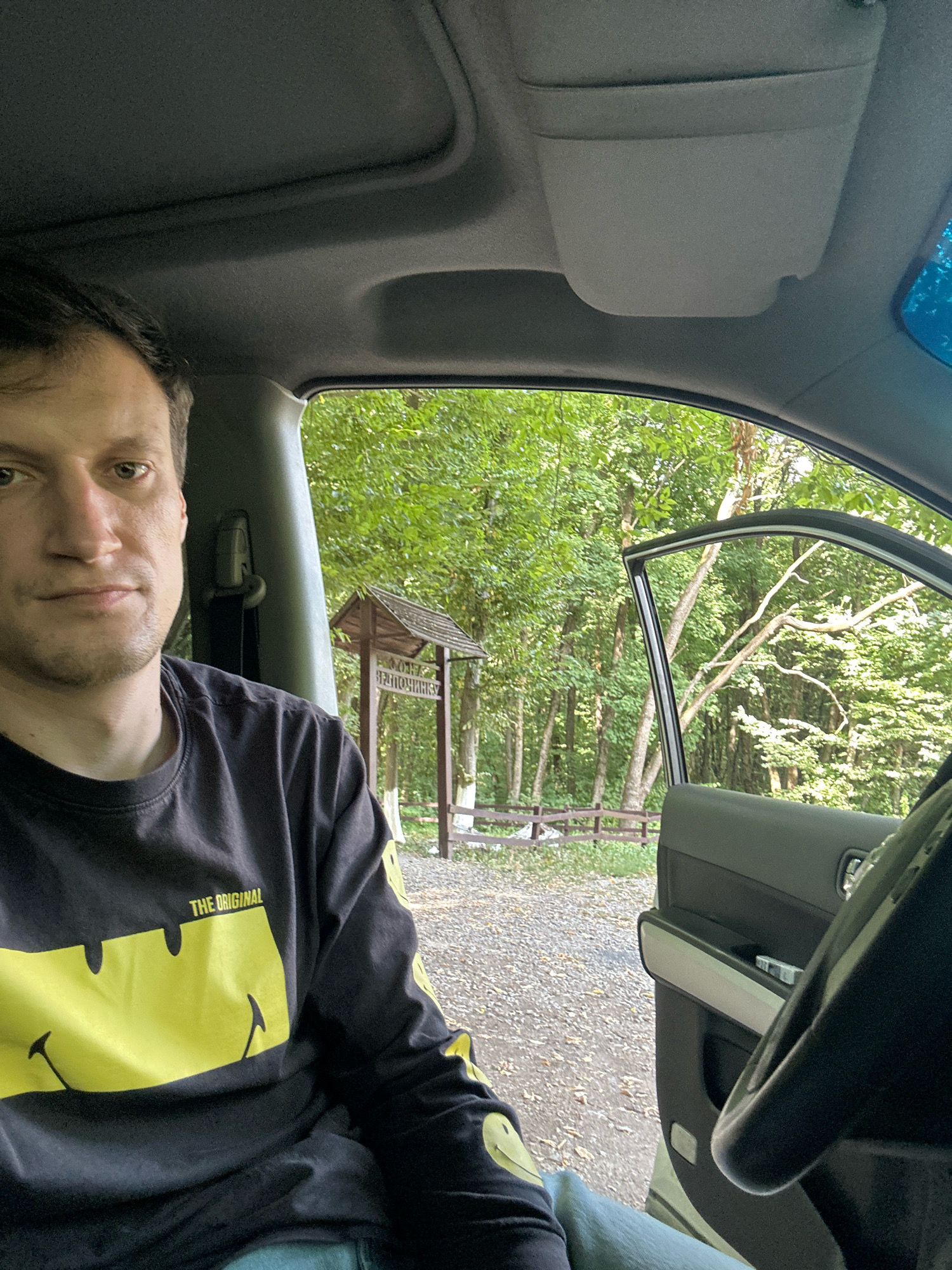 A selfie-style, eye-level shot shows a man sitting in the driver's seat of a vehicle. He is wearing a black long-sleeved shirt with a large yellow smiley face design and the text "THE ORIGINAL". The car door is open, revealing a wooded area with trees and a small wooden structure that appears to be a signpost. The ground outside the vehicle is covered with gravel. The man's left arm and shoulder are visible in the foreground, and the steering wheel is on the right side of the frame. The scene is set in the city of Shatava, Ukraine.