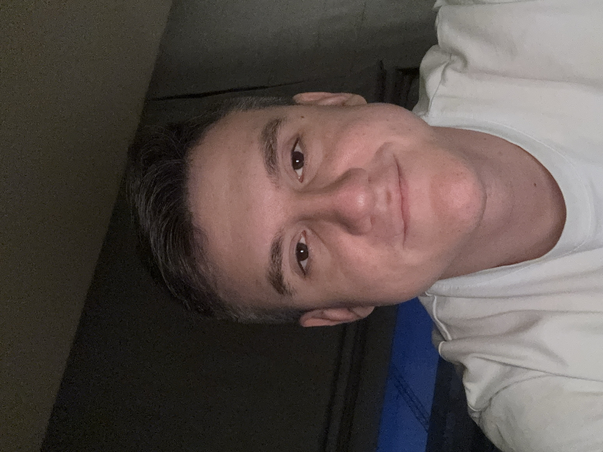 A man is lying down, likely indoors, with his head towards the top left of the frame. He has short, graying hair, and is wearing a white t-shirt. His face is turned towards the camera, displaying his eyes, nose, and mouth. Part of a dark wall or surface is visible in the upper left corner. A blue object or fabric is visible in the lower right. The location cannot be determined from the image.