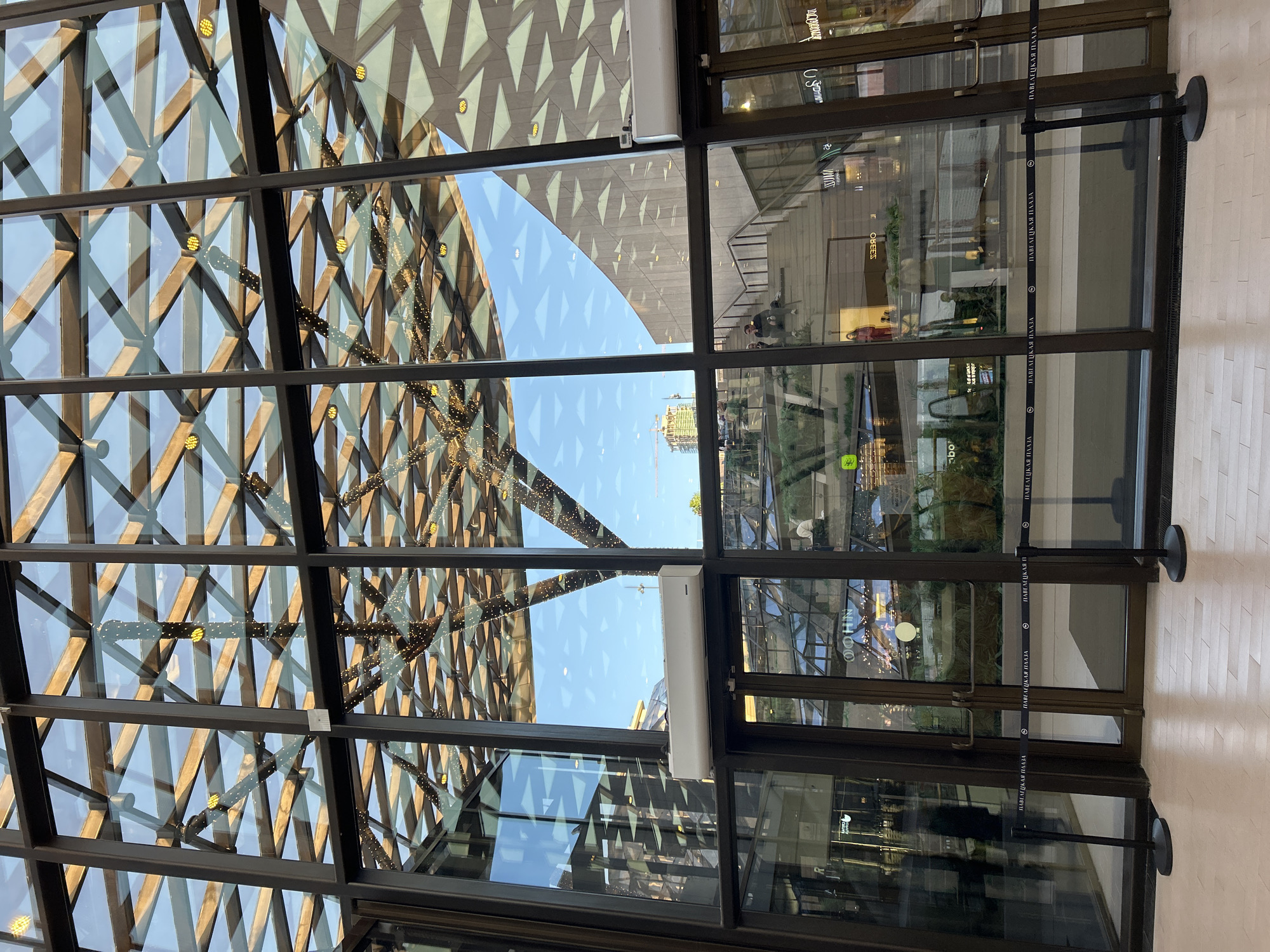 The image depicts a contemporary indoor space with a large glass facade, offering a view of the interior architecture and exterior sky. The interior features a wooden lattice ceiling adorned with string lights, creating a festive atmosphere. Below the ceiling, a multi-level structure with stairs is visible, and several people are seated or walking on the stairs. The lower part of the facade shows glass doors with black stanchions and velvet ropes in front of them, indicating a controlled entrance. Storefronts with illuminated signage are visible through the glass. A white air conditioning unit is mounted on the wall above the doors. The overall environment appears to be a modern shopping mall or public atrium.