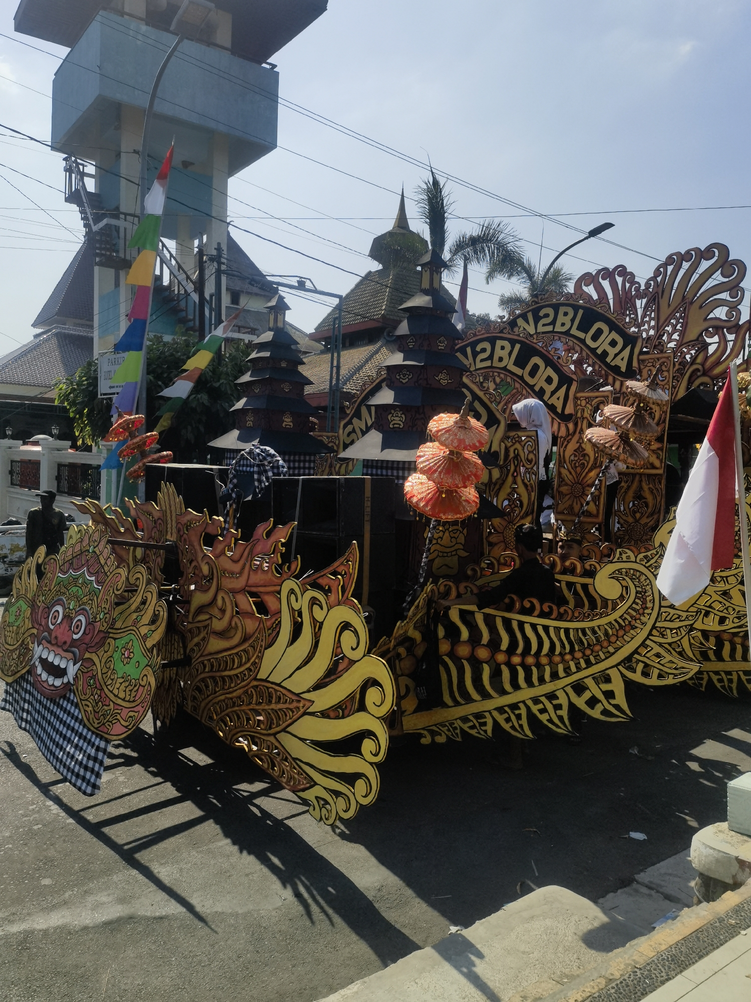 The image captures a parade float in Blora, Indonesia, likely for a cultural event or celebration. The float is elaborately decorated with gold and black ornate carvings, resembling mythological creatures or traditional Balinese art. A prominent banner across the float reads "N2BLORA". Several people are visible on and around the float, some appearing to be participants or organizers. In the background, traditional Balinese-style temples with tiered roofs are visible, along with a tall, light blue tower structure. Indonesian flags and colorful fabric banners are also present, adding to the festive atmosphere. The scene is set on a street under a clear blue sky, with shadows cast by the float and surrounding structures indicating it is daytime.