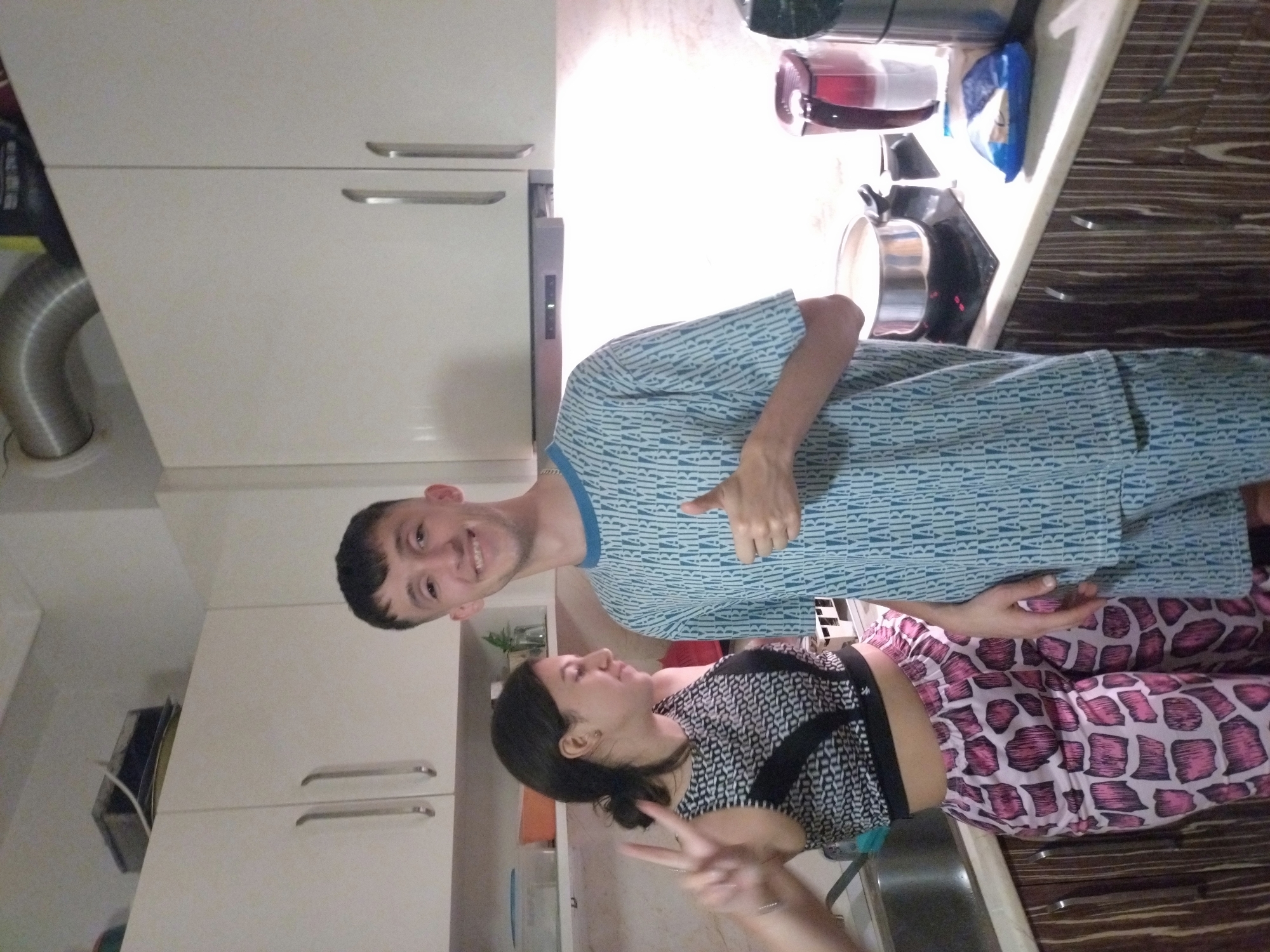 A photograph depicts two people in a kitchen environment. One person, possibly male, is wearing a blue patterned top and giving a thumbs-up gesture. The other person, likely female, is wearing a patterned top and pink patterned leggings, and making a peace sign with her hand. The kitchen includes white cabinets, a stainless steel sink, a kettle, and what appears to be a pack of crisps. The wooden-style floorboards can be seen. The location is presumed to be a private residence in Stara Zagora, Bulgaria, based on the prompt's information.