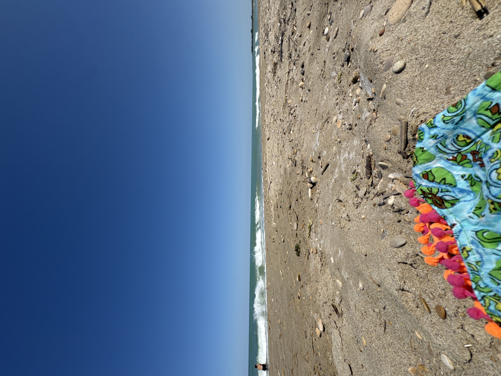 The image depicts a beach scene in San Benedetto del Tronto, Italy. The foreground features a close-up of sand with a patterned cloth adorned with pom-poms in shades of orange and pink. The sand is interspersed with small pebbles and driftwood. In the middle ground, the turquoise sea meets the sandy shore with gentle waves. A person is partially visible wading in the water on the left side of the frame. The background is dominated by a clear, vibrant blue sky.