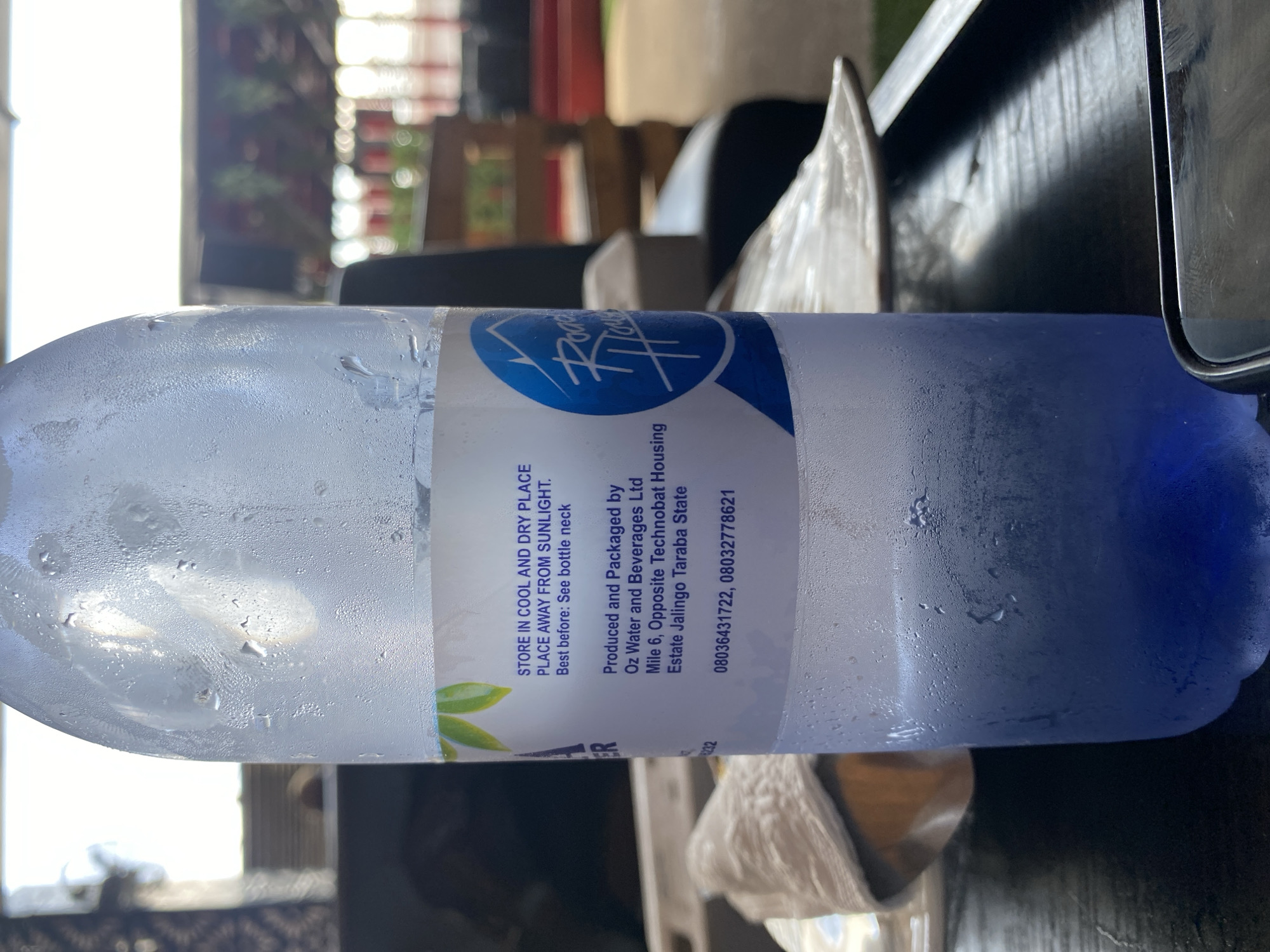 A condensation-covered plastic bottle of water stands on a dark wooden table. The bottle label is white and features green leaves at the top, followed by text and a blue logo with "Road House" written in white script. The text on the label reads: "STORE IN COOL AND DRY PLACE. PLACE AWAY FROM SUNLIGHT. Best before: See bottle neck. Produced and Packaged by Oz Water and Beverages Ltd Mile 6, Opposite Technobat Housing Estate Jalingo Taraba State. 08036431722, 08032778621". A portion of a white napkin or paper is visible to the left of the bottle, and further back, blurred figures and outdoor seating are discernible. The background suggests an outdoor or semi-outdoor seating area, potentially a restaurant or cafe, with a bright, hazy sky. The overall impression is a casual outdoor setting in a city, possibly a commercial area, given the street address information on the bottle.