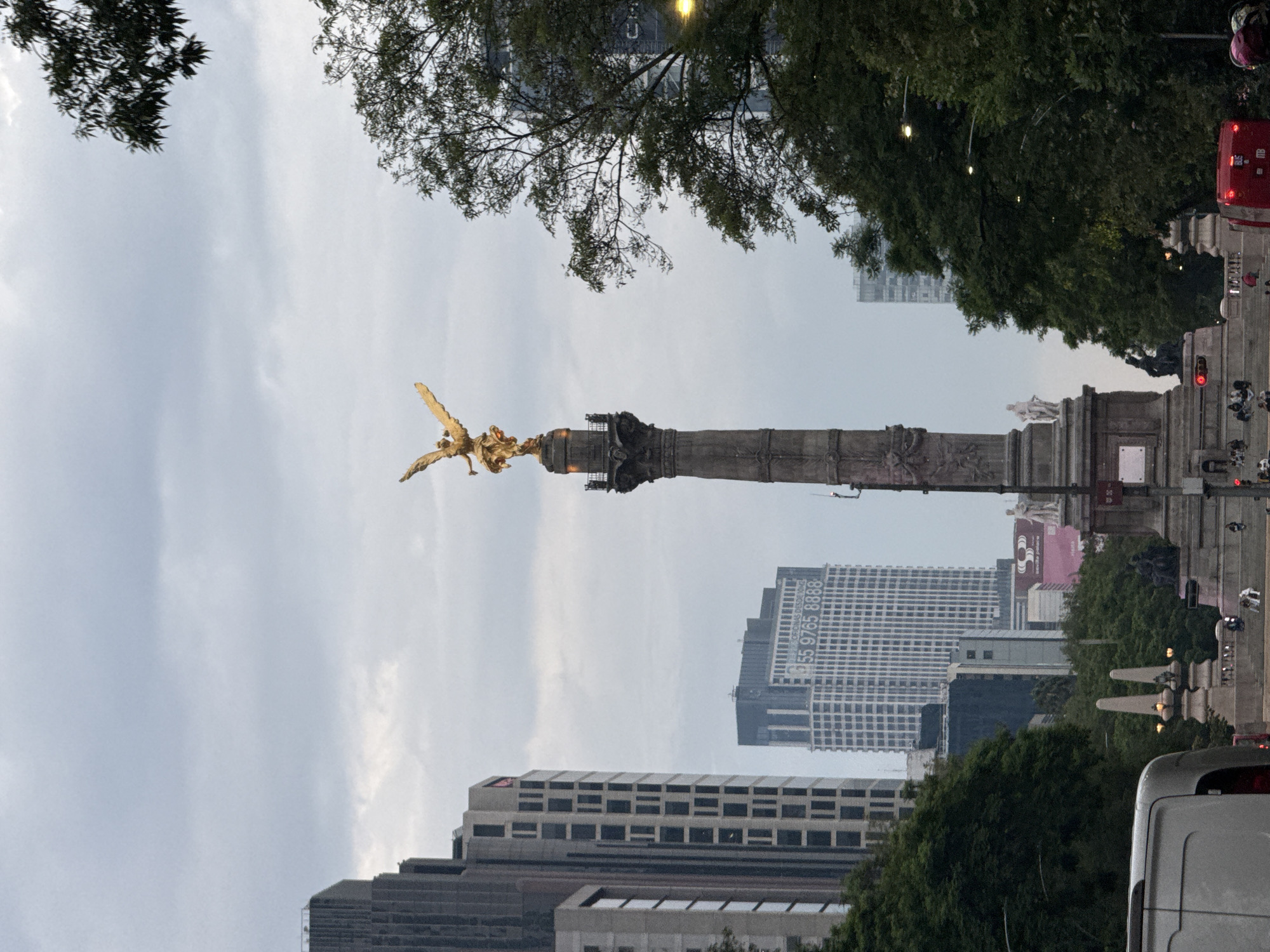 The image depicts the Angel of Independence monument in Mexico City, Mexico, against a backdrop of cloudy skies and various buildings. The monument is centrally positioned, with the golden angel figure prominently visible. The surrounding urban environment includes a mix of modern skyscrapers and older architectural styles. There are trees on either side, partially obscuring the view, along with traffic and urban infrastructure. The composition suggests a street-level perspective, possibly captured from a moving vehicle or pedestrian viewpoint. The monument appears well-maintained, and there are signs of urban activity.