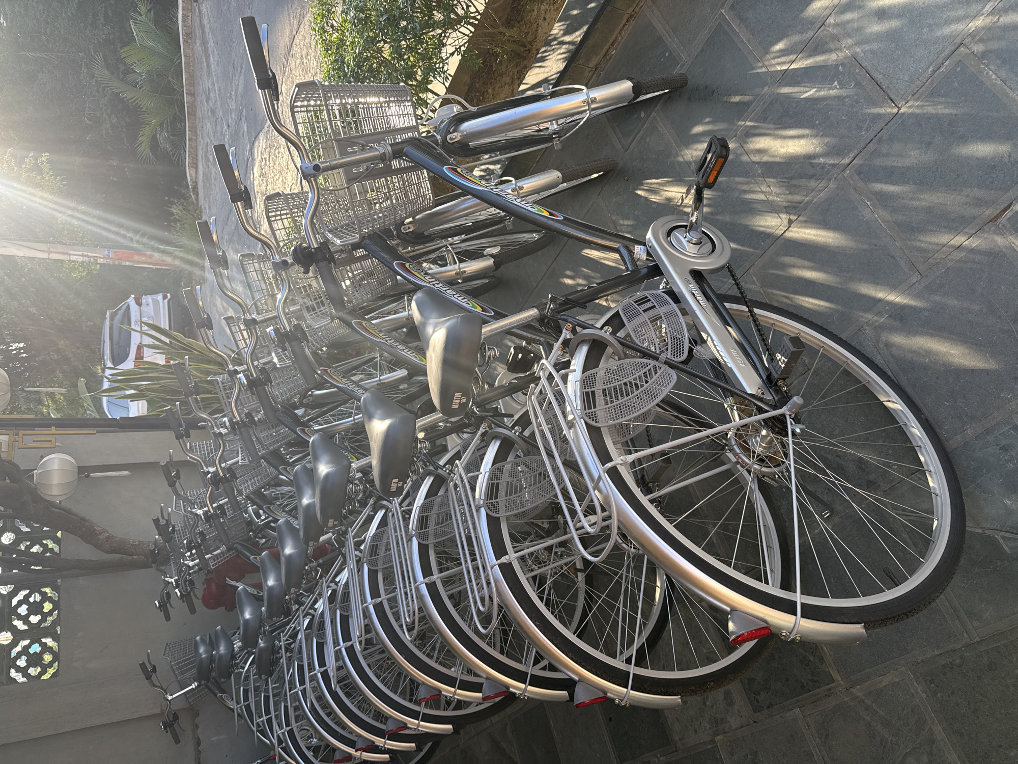 A horizontal shot displays a row of bicycles parked on a gray, paved surface. The bikes are silver and black, each equipped with a basket in front and a seat. The brand "Martin" is visible on the seats. The bikes are lined up closely, their wheels overlapping. Behind the bikes, greenery is visible along with a glimpse of a white vehicle. The sun is shining brightly, creating lens flare. The location is in Hội An Ward, Vietnam.