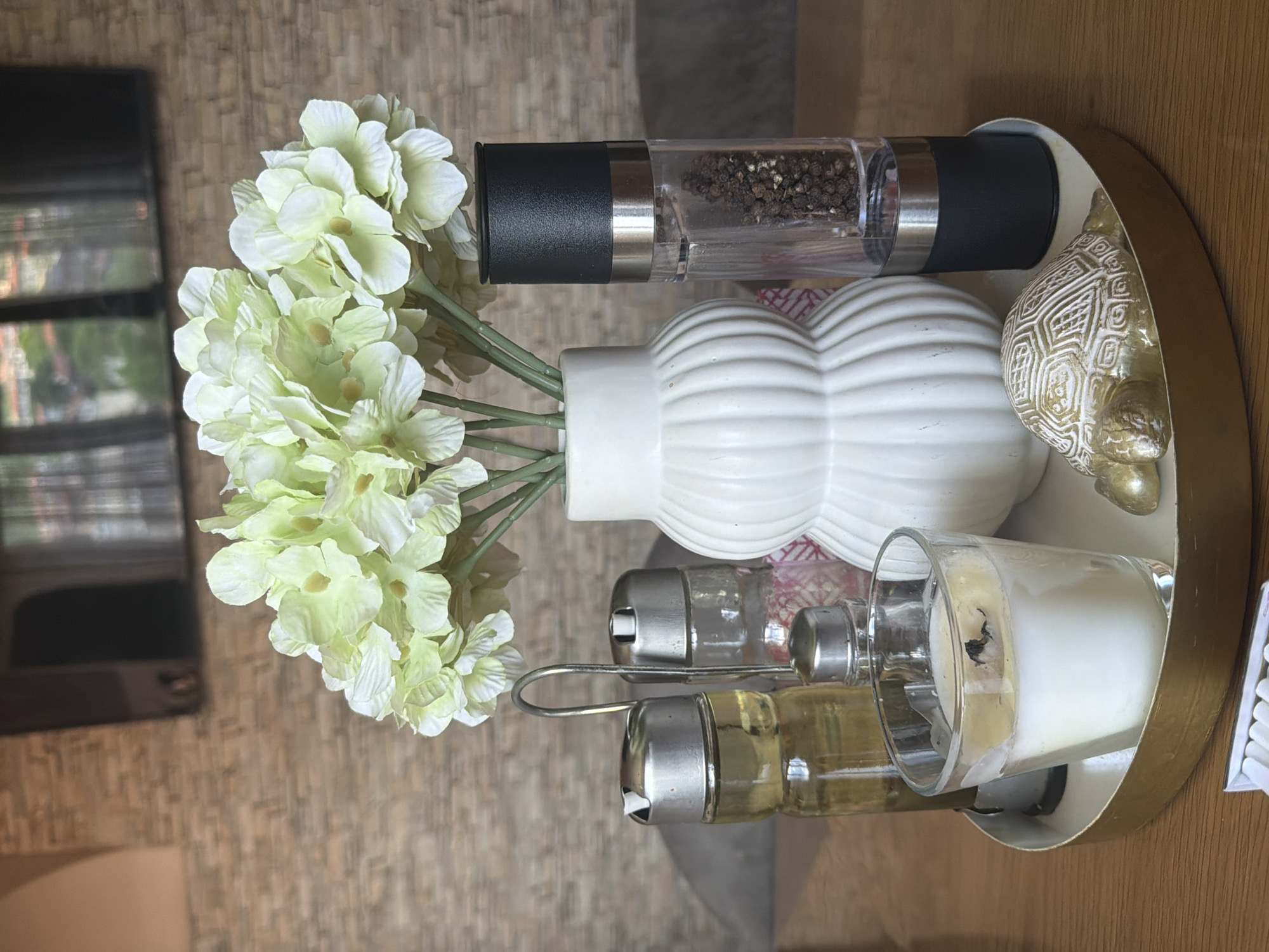 An overhead shot, rotated ninety degrees clockwise, captures a decorative arrangement on a round, golden-rimmed tray. At the center left, a white ribbed vase holds a bouquet of light green and white artificial hydrangeas. To its right, a tall, clear pepper grinder, filled with black peppercorns, stands upright with a black and silver top.
Next to the pepper grinder on the right, a small golden figurine resembling a turtle with an intricately patterned shell rests on the tray. In front of the vase and pepper grinder, a metal caddy holds two small glass shakers with silver tops; one contains white crystals (likely salt), and the other holds a yellowish liquid or substance.
To the far right, a white candle in a clear glass holder is partially visible, showing a burned wick. A small white box, possibly containing matches, is also partially seen on the tray's edge. The tray rests on a light brown wooden surface. In the background, a dark, rectangular reflection (possibly from a window or screen) shows blurry shapes that suggest buildings or an outdoor scene. There is no visible text. The location is Shumen, Bulgaria, though no specific visual cues confirm this in the image.