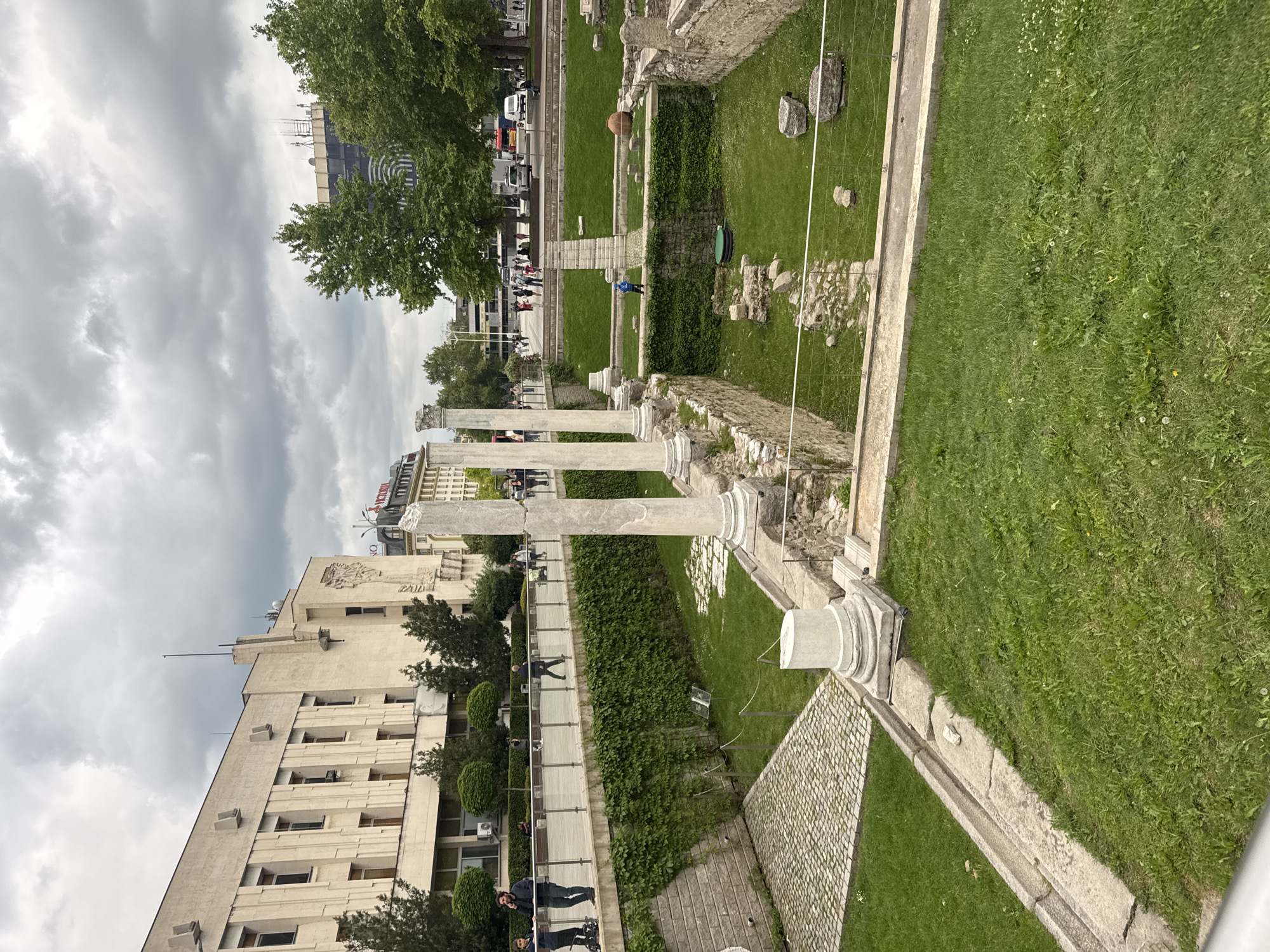 The image depicts the Ancient Stadium of Philippopolis in Plovdiv, Bulgaria. The stadium's remaining columns stand amidst grassy areas, separated by short, clipped hedges. A paved walkway runs parallel to the stadium, with people strolling along it. Adjacent to the walkway is a large, multi-story building with numerous windows. The sky is overcast with heavy clouds. The general scene presents a blend of ancient ruins integrated into a modern urban environment.