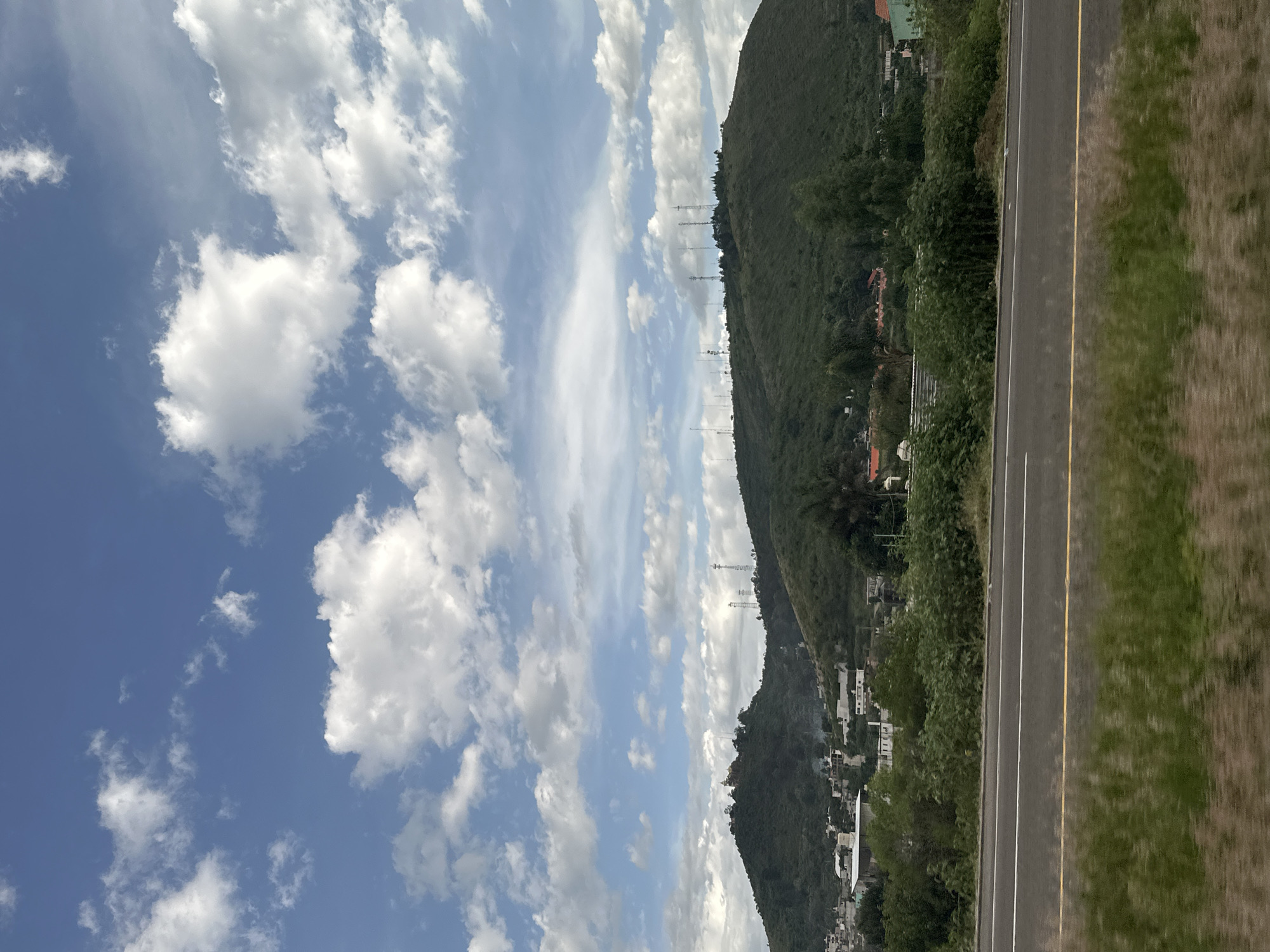 The image captures a wide view of a semi-urban landscape with prominent hills in the background, under a partly cloudy sky. The foreground features a paved road with distinct white and yellow lane markings, bordered by grassy verges. Beyond the road, a band of green foliage and scattered buildings can be seen, indicating a transition zone between the highway and the populated hillside.
The hills dominate the midground and background. The left hill is covered in dense green vegetation and has a cluster of buildings, possibly houses and smaller structures, nestled on its lower slopes. A prominent structure with a dome-like feature, possibly a church or a monument, is visible on the crest of this hill. To the right of this hill, the landscape slopes downward before rising again to a larger, more extensively populated hill. This second hill is also densely vegetated, with numerous telecommunication towers scattered across its summit and upper slopes. The structures on this hill appear to be a mix of buildings and what could be settlements.
The sky is a vivid blue, populated by scattered white cumulus clouds of varying sizes and densities. Some clouds are wispy and spread out, while others are more voluminous and distinct. The lighting suggests it is daytime, with the sun casting shadows that are not prominently visible, indicating diffused light, possibly due to the cloud cover.
The overall scene suggests a location on the outskirts of a city or town, where natural topography meets human development. The presence of numerous communication towers on the hills indicates a strategic location for broadcasting or telecommunications infrastructure. The combination of residential buildings, vegetation, and the highway points to the specific geographic and urban context of San Bernardino Chalchihuapan, Mexico, a place known for its hilly terrain and increasing development.