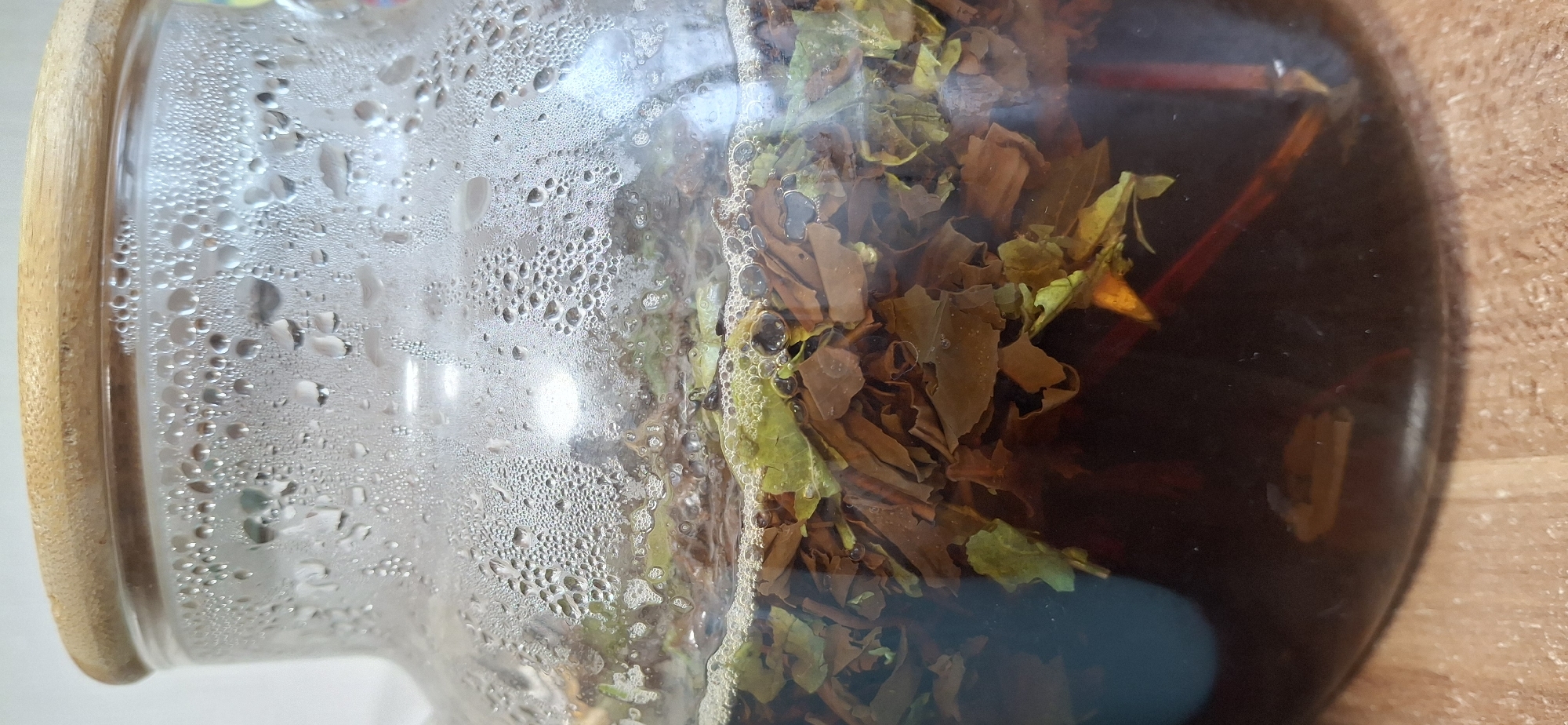 The image shows a close-up view of a glass jar or teapot, with a wooden rim around the top. The left side of the jar is covered in condensation, consisting of numerous small water droplets. The right side of the jar contains brewed tea, with loose tea leaves visible inside the dark liquid. Some of the tea leaves appear to be brown or green. There are also some reddish brown sticks visible. There are bubbles around the edges of the tea leaves. The background appears to be a wooden surface. The location of the image is unknown.