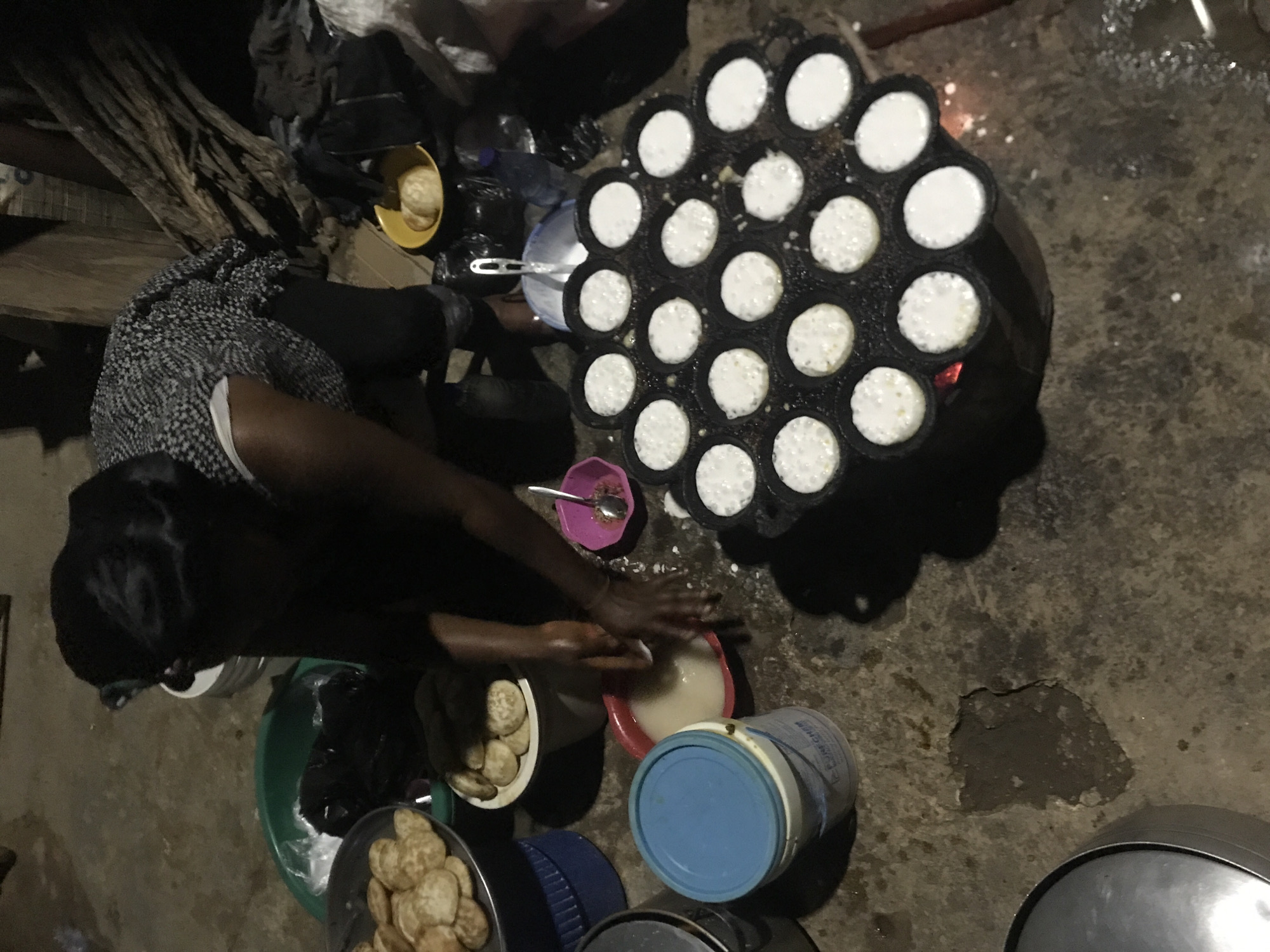 A person, likely a woman based on her hairstyle and clothing, is preparing food outdoors in New Stadium, Nigeria. She is bent over a cooking apparatus that appears to be a traditional stove with a dark, round cooking pan filled with small, circular indentations. Each indentation contains a white, liquid batter, suggesting she is making pancakes or a similar type of fritter. Her hands are in a red bowl filled with a white liquid, possibly a part of the batter preparation.
Around her, various containers and food items are visible. To her left, a stack of golden-brown, flattened fried dough items are piled on a grey plate, which is itself placed on a blue bucket. Next to this, a green basin holds some items covered by a black bag. Further to the right, a yellow bowl contains a few more of the fried dough items. There are also other buckets and bowls scattered around the ground, which appears to be a dirt or concrete surface with some debris. In the background, what looks like dried wood or logs are propped up. The lighting suggests it is either dusk or night, with a strong focus on the immediate cooking area, likely illuminated by a lamp or fire.