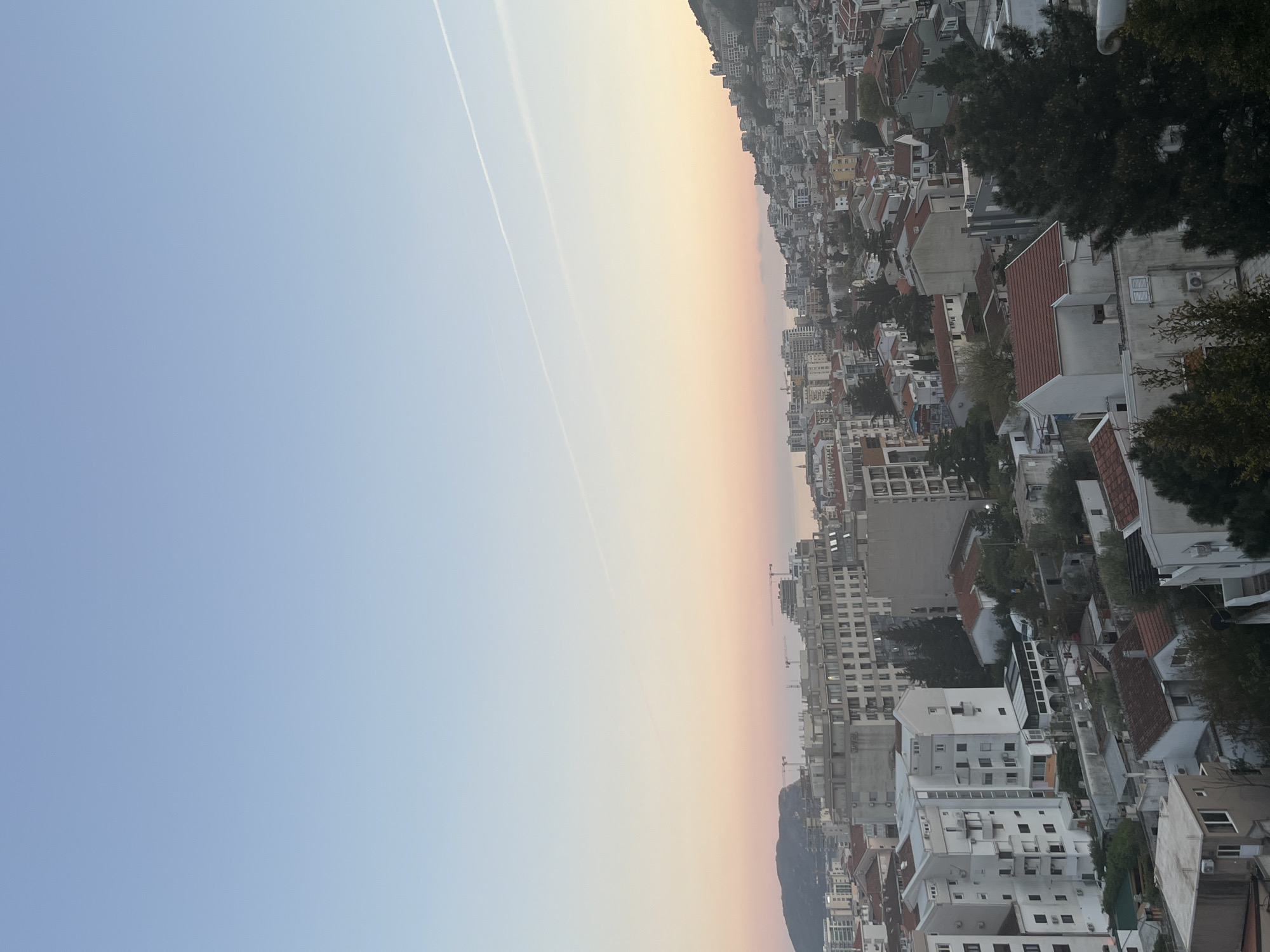 The image shows a portion of the city of Budva, Montenegro, taken from an elevated perspective, possibly a hillside or a tall building. The cityscape is densely packed with buildings of various sizes and designs. Red-tiled roofs are interspersed among white and grey modern structures. Trees are scattered among the buildings. In the background, there is a gradient sky fading from blue to orange, indicating either sunrise or sunset. There are two contrails in the sky. The image is oriented vertically.