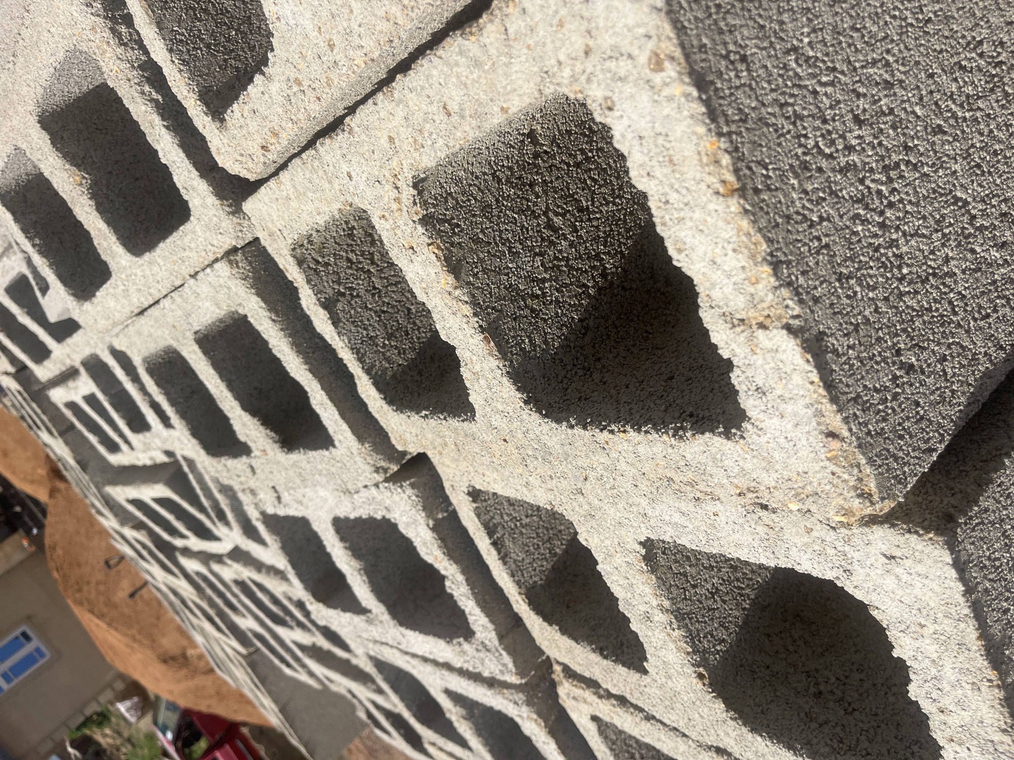The image shows a close-up view of stacked concrete cinder blocks, casting long shadows. The blocks have a rough, porous texture. The lighting suggests it is daytime, with harsh sunlight creating strong contrasts between the illuminated surfaces and the shadowed openings of the blocks. In the background, partially visible, is a building with a blue-framed window, and some vegetation. The presence of a red vehicle in the background, along with the building, suggests a residential or construction setting. The scene is outdoors, likely in an area where construction is taking place or materials are stored. No people are visible in the image. There is no visible text.