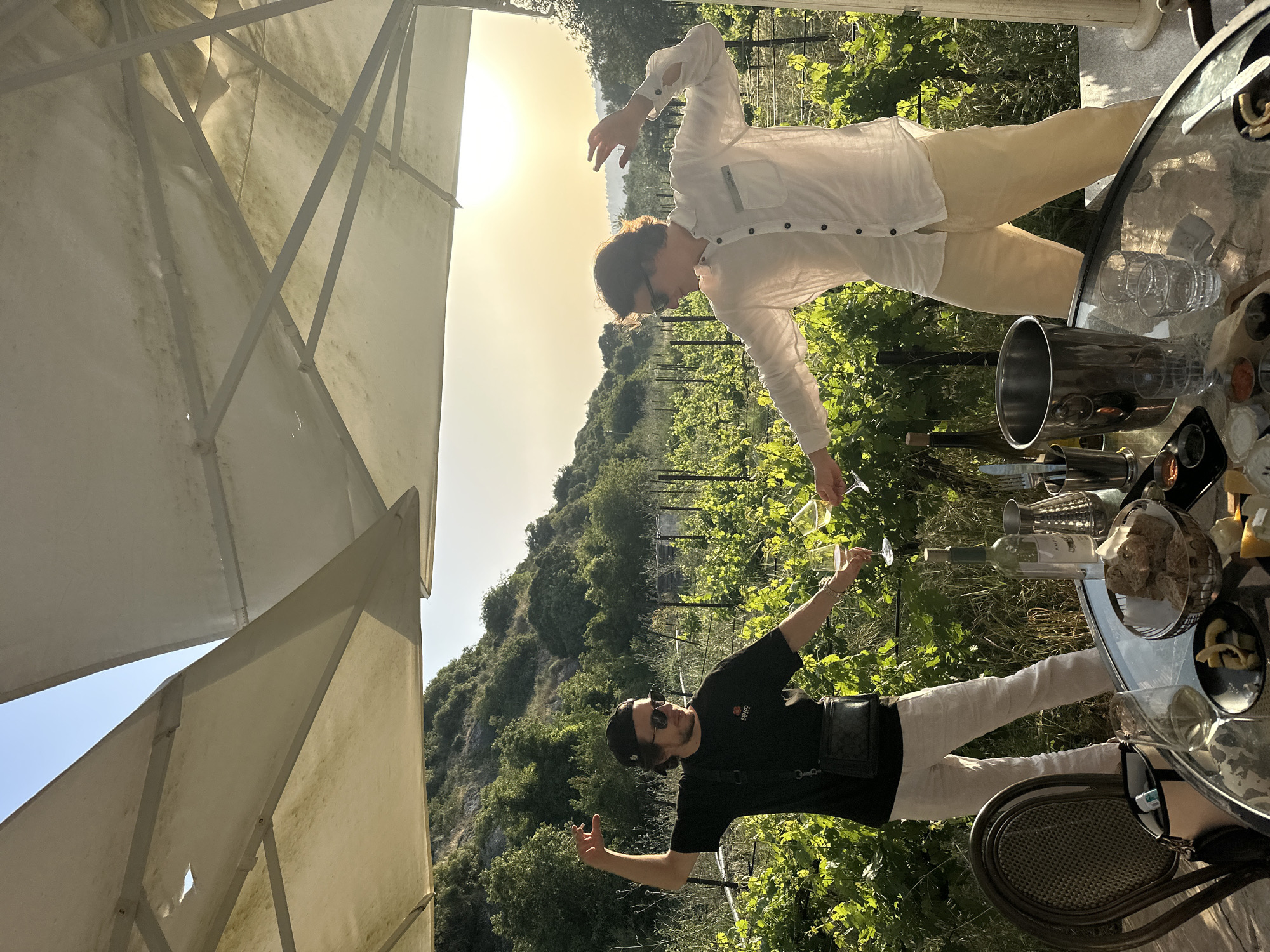 Two people stand outside next to a glass table with wine glasses and bottles in Hof HaCarmel Regional Council, Israel. One person wears a black shirt and a baseball cap and raises one arm into the air. The other person wears a white button-down shirt and yellow pants and has their arms raised as well. Both people are in a vineyard. There is a mountain with trees and greenery visible in the background and a sun behind them.