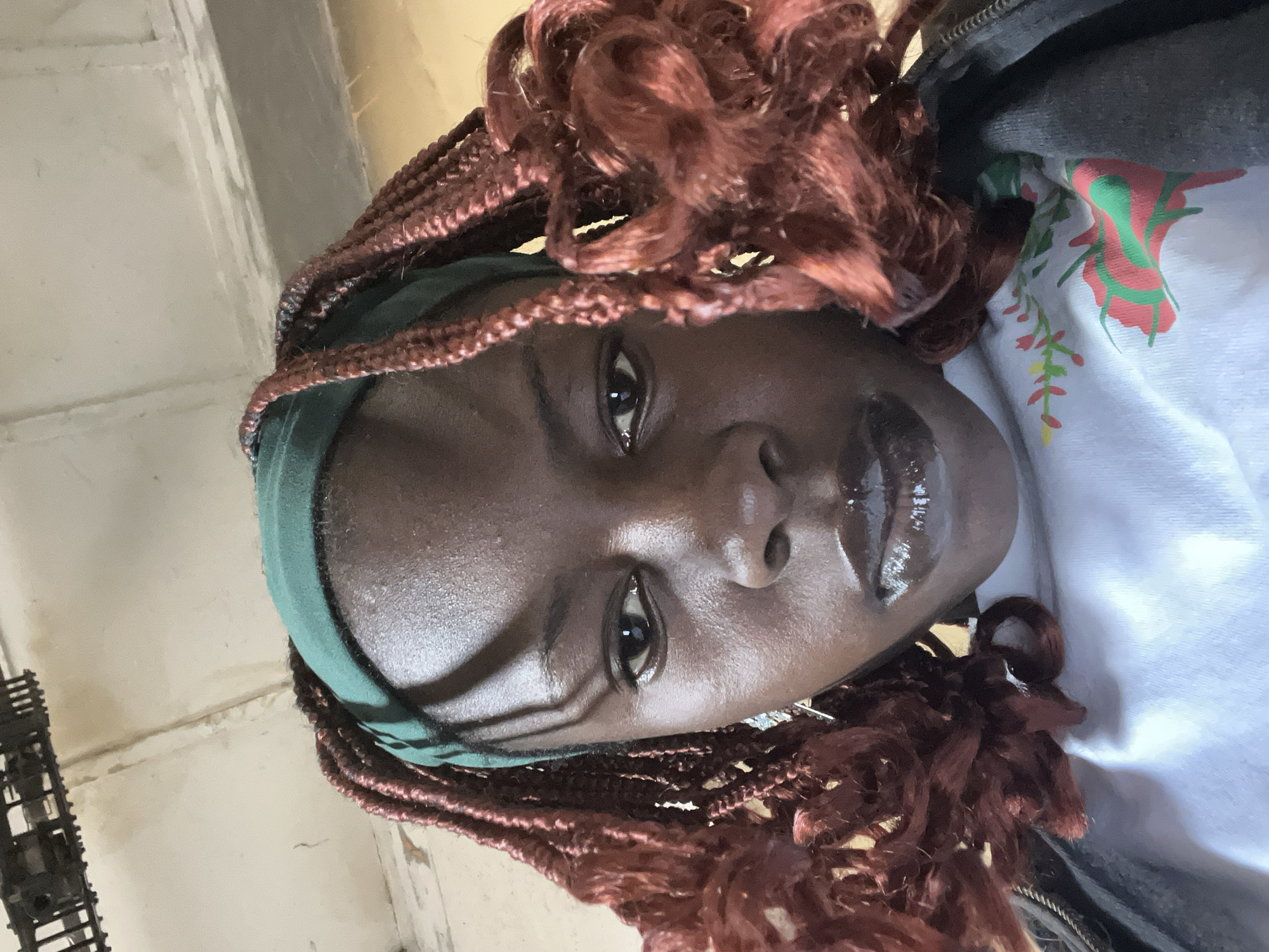 The image shows a close-up, low-angle selfie of a young Black woman indoors, looking down at the camera. She has dark skin, braided auburn hair, and is wearing a green headscarf. Her lips are glossy and dark. A white t-shirt with a floral design is visible beneath a grey zipped jacket. The background appears to be a ceiling with some fixtures, and a wall with staining. The overall impression is that of a casual, personal photograph. There is no information in the image to suggest it was taken in Jos, Nigeria.