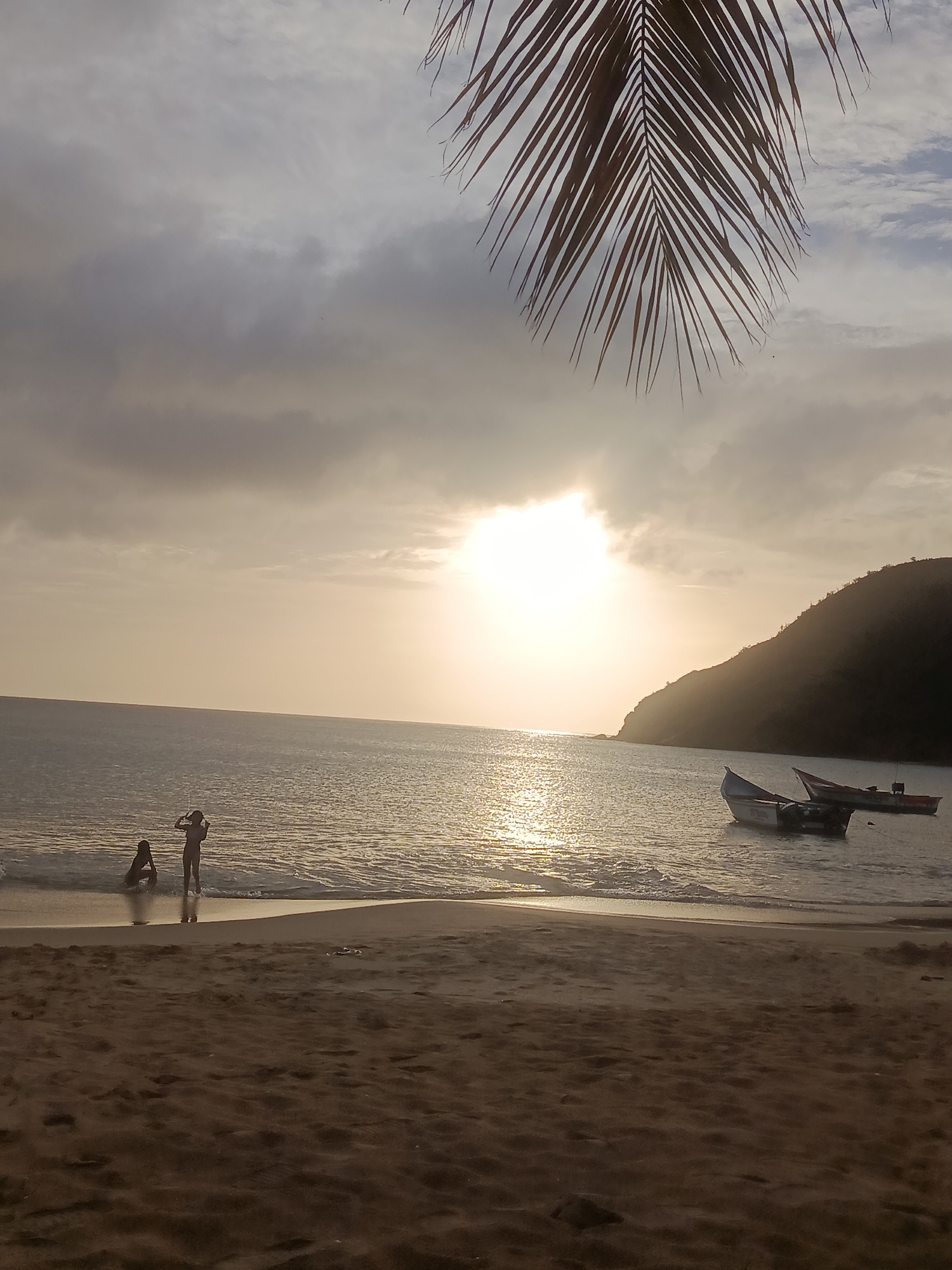 The image shows a beach scene at sunset in Juan Griego, Venezuela. The sun is low on the horizon, casting a warm glow over the water and sky. Two people are in the shallow water near the shore, one appearing to be sitting and the other standing. Several boats are anchored in the water, with a hilly coastline visible in the background. A palm frond hangs from the top of the frame, obscuring part of the sky. The sand in the foreground is dark and textured, with some small objects visible on it. The overall mood is tranquil and serene.