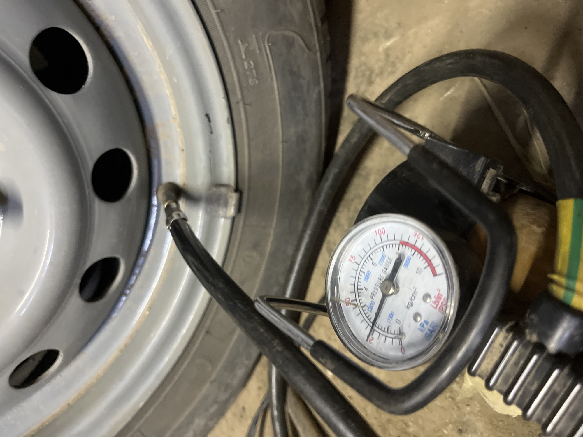 The image shows a close-up view of a car tire and a tire pressure gauge. The tire is grey and has several circular cutouts. A black hose with a metal nozzle is attached to the tire's valve stem. A circular tire pressure gauge with a white face and black markings is connected to the hose. The gauge displays readings in psi, kg/cm², and bar. The background appears to be a concrete floor. The scene is likely located in Krasnyye Barrikady, Russia, where this activity of checking tire pressure would be performed.