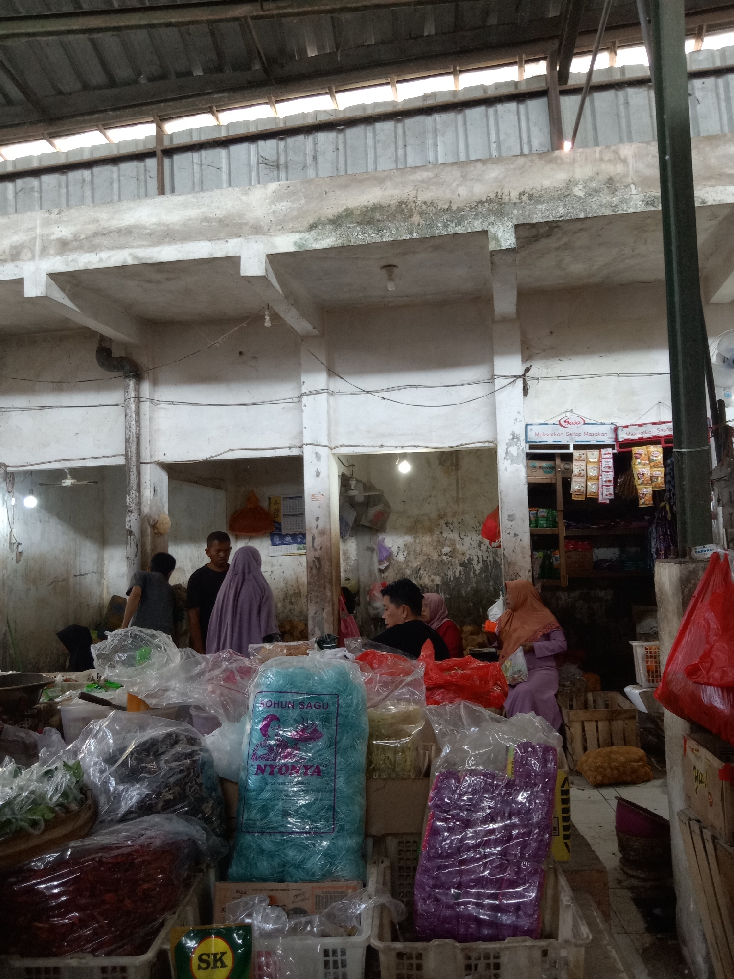 The image depicts an indoor market scene in Sugihwaras, Indonesia. The market appears to be a general goods or food market, with stalls selling various packaged food items, produce, and other commodities. The structure is utilitarian, with exposed concrete columns and beams, a corrugated metal roof, and open-air sections allowing natural light.
In the foreground, several large plastic-wrapped packages are visible. One prominent package is a large, translucent blue bag filled with what appears to be rice vermicelli or a similar noodle product, labeled "SOHUN SAGU NYONYA". To its right, a purple package, also plastic-wrapped, contains a similar product. To the left, a large red pile of what looks like dried chili peppers is partially visible in a white plastic basket. Other plastic-wrapped packages containing green leafy vegetables and dark dried goods are also present.
Several people are observed in the market. In the mid-ground, a man in a black t-shirt is speaking with a woman wearing a light purple headscarf and a black garment. To their left, another person in a dark grey t-shirt is partially visible. Further back, a woman in an orange headscarf and a purple dress is interacting with someone off-camera. In the background, a shelf unit stocked with various food packets and seasonings is visible, with a sign that reads "Sasa Melezatkan Setiap Masakan". A calendar is also hanging on the wall. The environment is somewhat cluttered, with goods displayed on the floor and on shelves, indicative of a busy marketplace. A fan is visible in the background on the right.