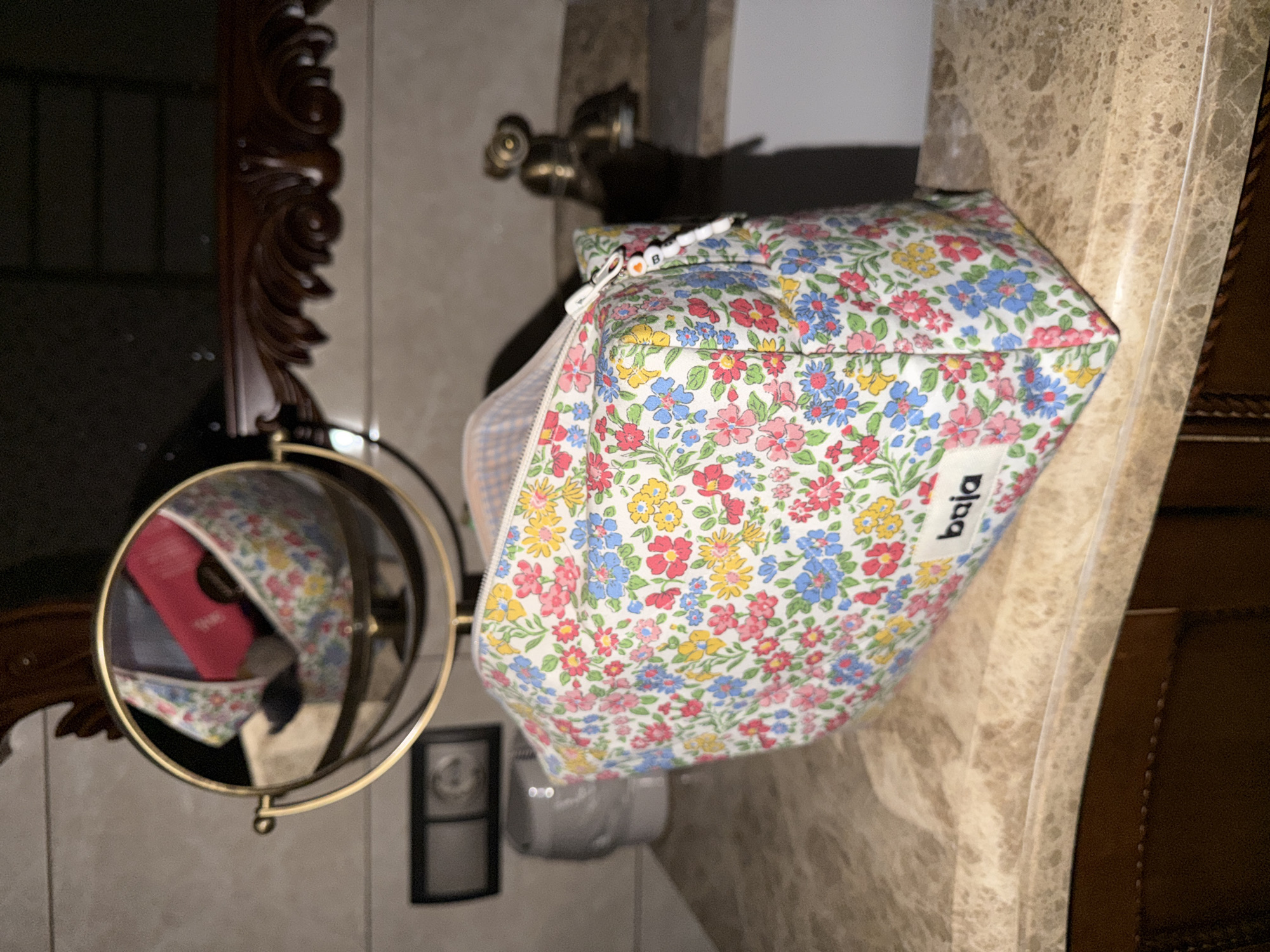 A floral patterned cosmetic bag is resting on a marble countertop in a bathroom setting. The bag features a white label with the word "Baja" printed on it. In the background is a vintage-style mirror with an ornate frame and a small magnifying mirror attached to it. The reflection in the mirror reveals a partially visible red object inside the bag. Behind the bag is a faucet and other bathroom fixtures. A switch on the wall is also visible. The environment is indoors, suggesting a private residence. The specific location of Mishutkino, Russia, cannot be determined from the image alone.