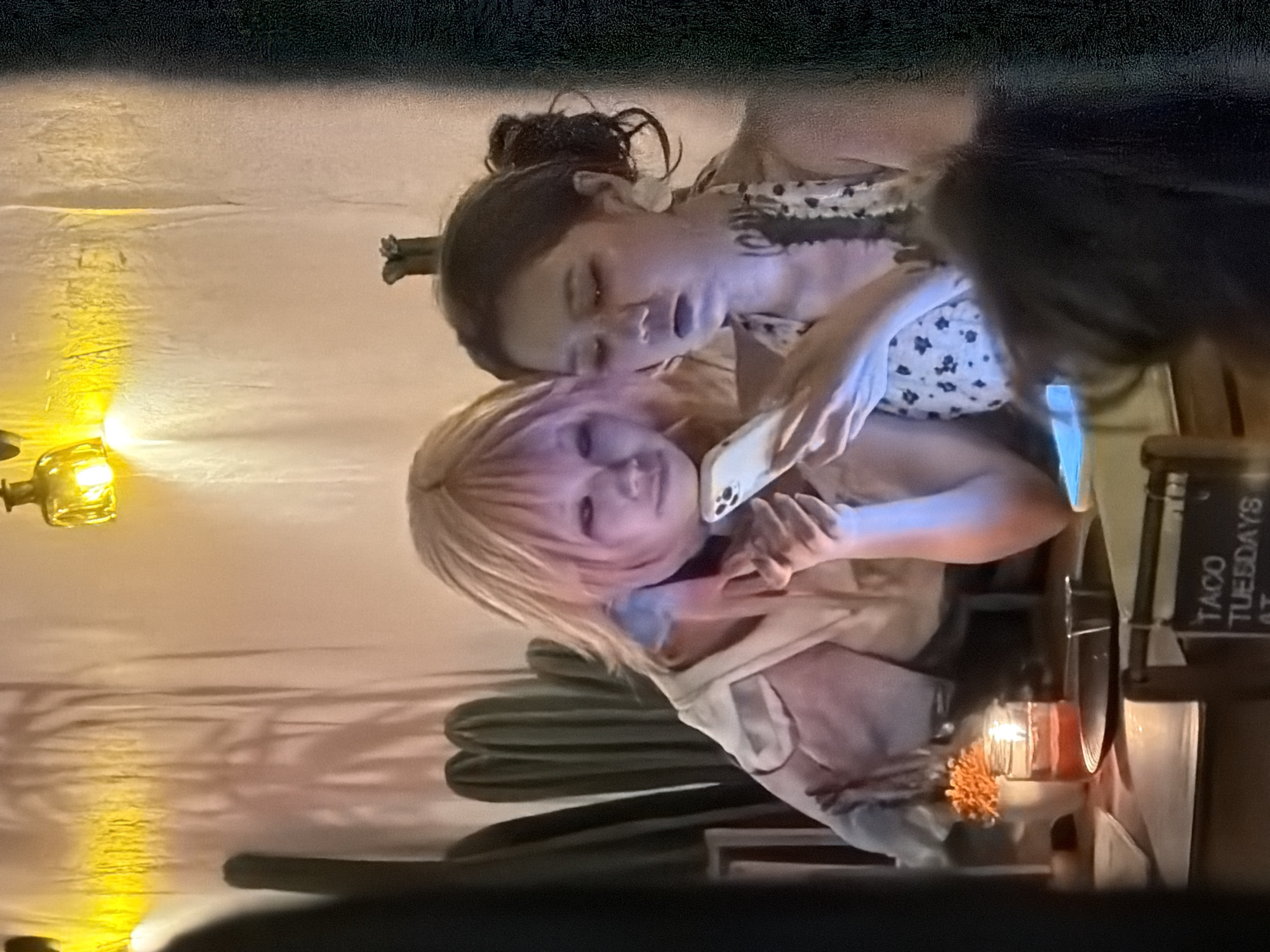 Two individuals are seated at a table with a lit candle and a potted plant. The person on the left has pink hair and is looking at a smartphone. The person on the right, with dark hair pulled back, is also looking at the smartphone. The background features a textured wall with lighting and large, dark, cactus-like figures. The lighting in the scene is warm, with a noticeable blue cast on the faces of the individuals due to the smartphone's screen. The environment appears to be an indoor setting, possibly a restaurant or bar, given the table and seating arrangement. The "Taco Tuesdays" sign partially visible in the foreground suggests a specific day of the week and a potential food establishment. The location is identified as North Kuta, Indonesia.