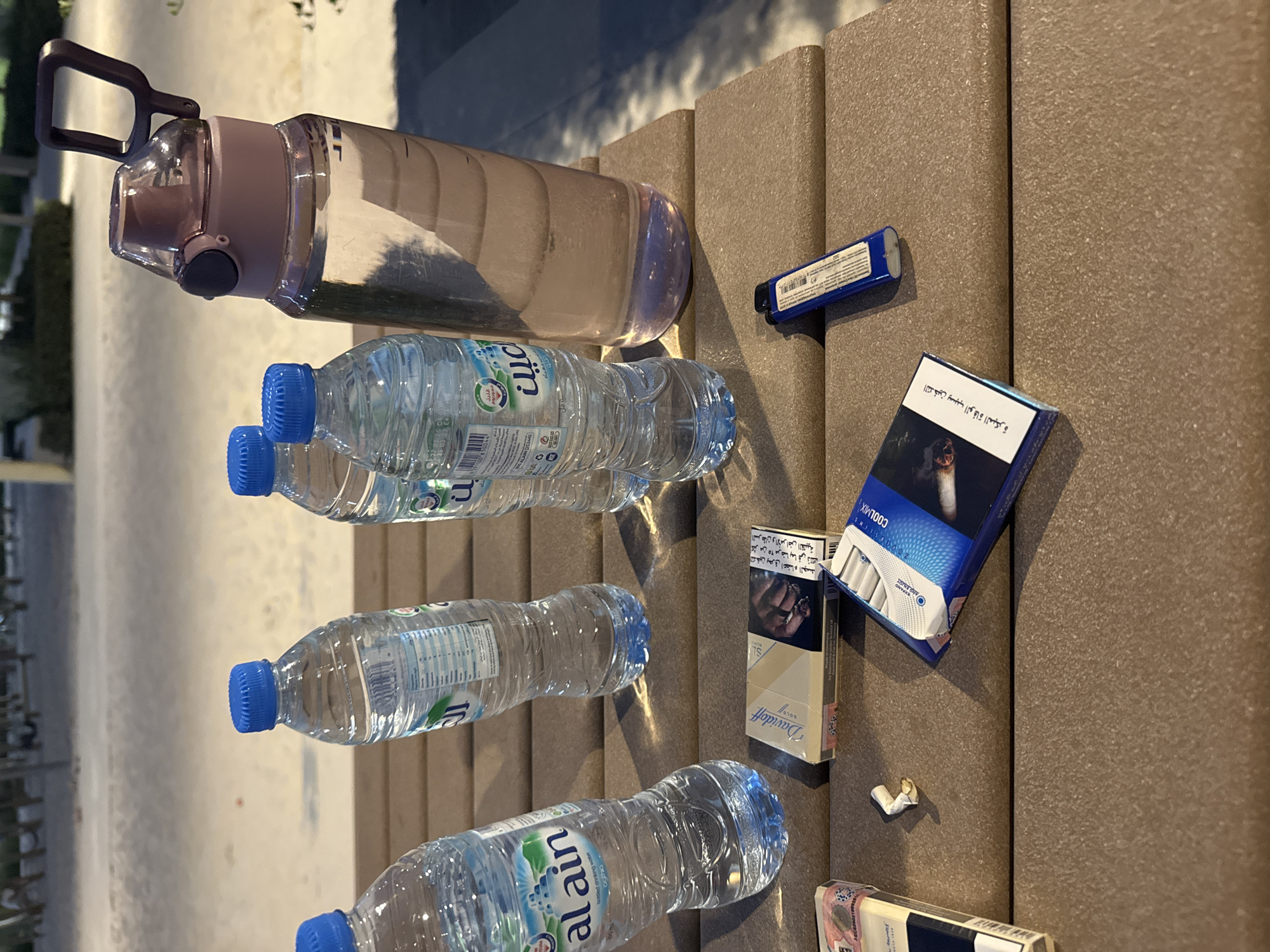 The image displays a collection of items placed on a textured, brownish outdoor surface, likely a bench or table. The foreground features a large, clear reusable water bottle with a pink cap and dark handle, three smaller disposable plastic bottles of Al Ain bottled drinking water, two cigarette packs, a blue lighter, and a discarded cigarette butt.
The setting appears to be outdoors during daytime, indicated by the bright lighting and the blurred background showing a paved area and some greenery. The Al Ain water bottles and the Arabic text on the cigarette packs are strong cues for the Middle Eastern location, consistent with Dubai, United Arab Emirates.
The light-colored cigarette pack is identified as "Davidoff GOLD SLIMS" and carries an Arabic health warning: "التدخين يحرق اعضاء الجسد / أكثر من ٢٥ مرضا بما في ذلك / السرطان والأمراض القلبية" (Smoking burns body organs / more than 25 diseases including / cancer and heart diseases). The blue cigarette pack, labeled "COOLMIX", displays another Arabic warning: "التدخين يسبب الوفاة المبكرة" (Smoking causes premature death). A blue lighter is also visible, with "ALAIN 25246" appearing in the OCR, possibly from a label on it. No specific activity is taking place, but the items suggest a break or leisure moment.