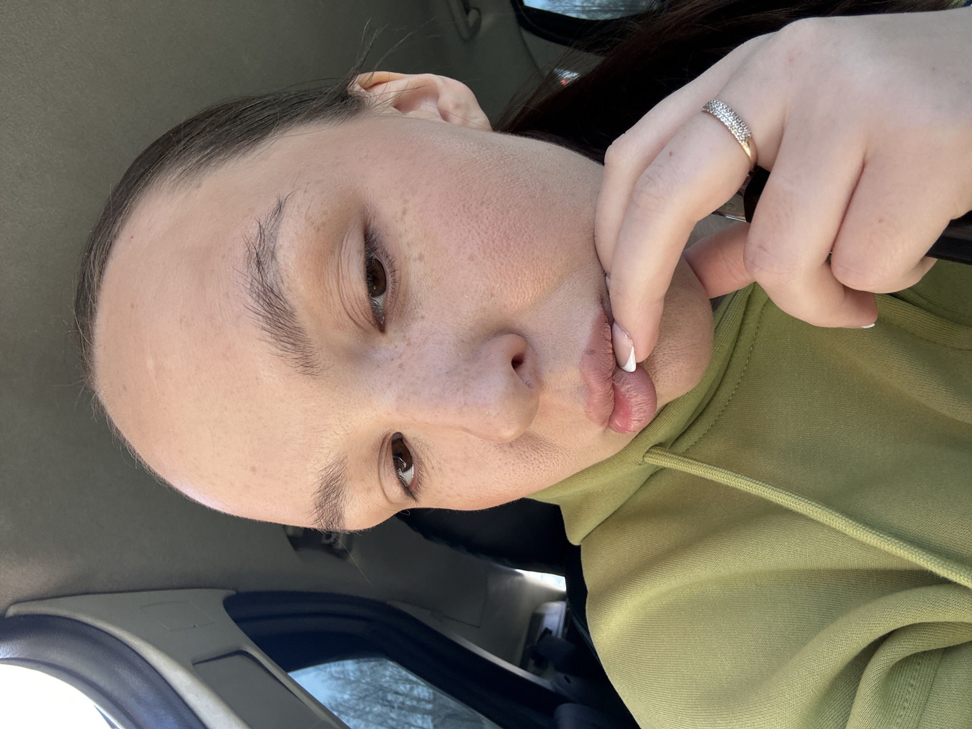 A selfie taken indoors, presumably inside a vehicle, featuring a light-skinned person with brown hair, wearing a light green hooded sweatshirt and a silver ring on the ring finger of their left hand. The person's face is slightly angled, showing brown eyes, freckles, and a pouting expression as they touch their lower lip with their index finger. Parts of the vehicle's interior are visible, including the gray roof lining and a glimpse of the window and outdoor scenery, which includes trees. The location in the city of Chaikovsky, Russia, is not determinable from the image.