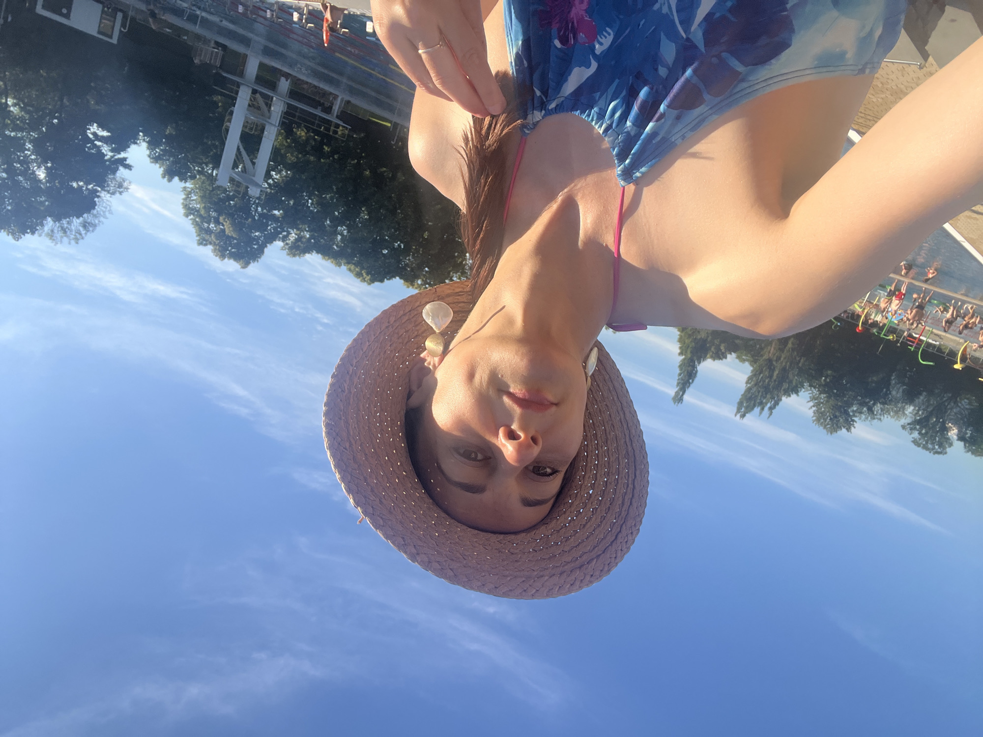 A woman wearing a straw hat and a blue floral swimsuit poses for a selfie outdoors. She has on large, circular earrings and a ring on her left hand. The background shows a swimming pool area with people, trees, and a diving board under a blue sky with wispy clouds.