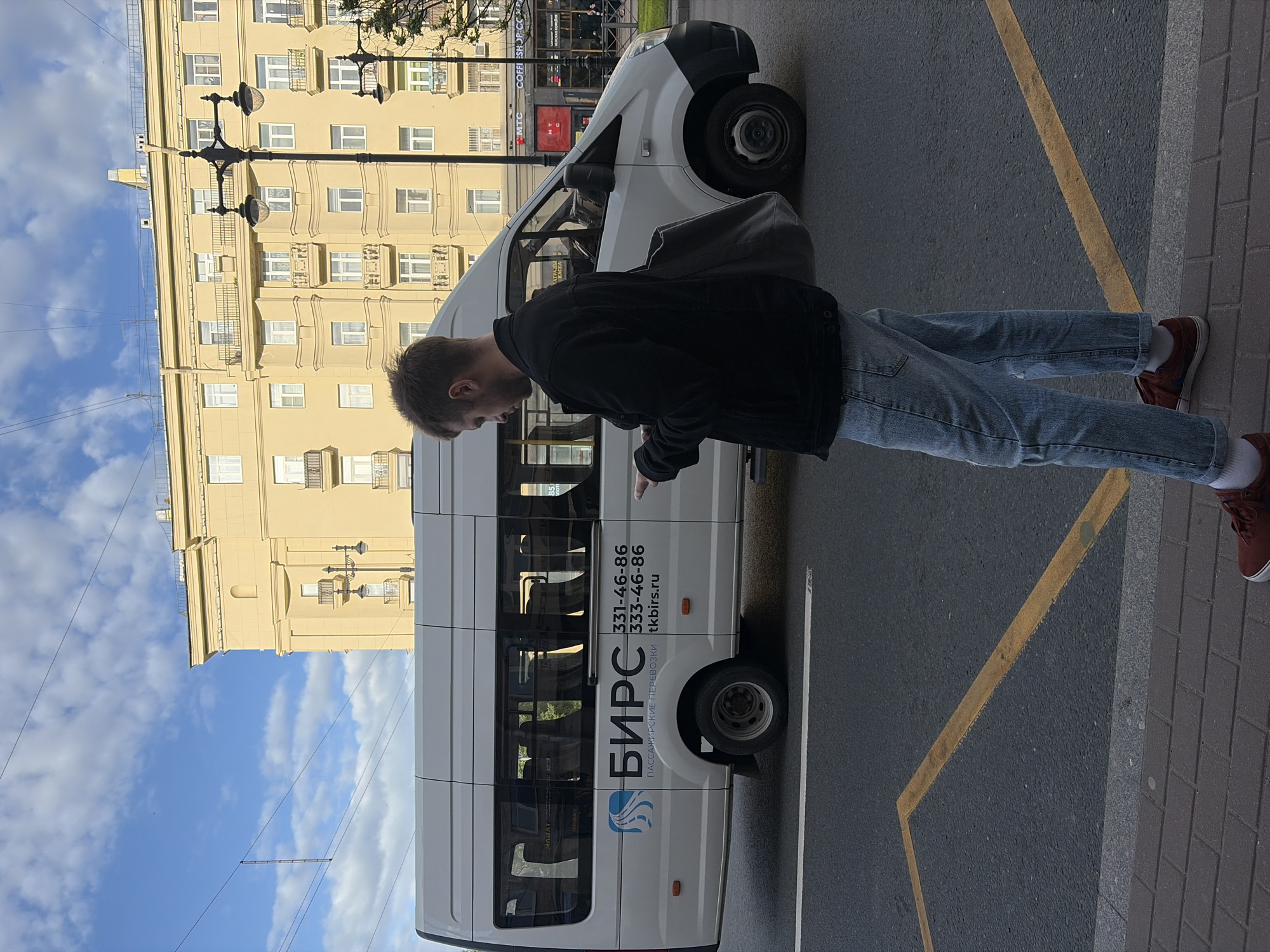 The image shows a man standing next to a white passenger van on a street in Saint Petersburg, Russia. He's wearing a black jacket, light-colored jeans, and reddish-brown shoes. He appears to be gesturing towards the van, which has the name "БИРС" and two phone numbers, "331-46-86" and "333-46-86," along with the website "tkbirs.ru" written on its side. The inscription on the van is in Russian and indicates the company is engaged in passenger transportation.
The van is parked next to a yellow painted line marking a designated zone on the asphalt street. Adjacent to the street is a sidewalk paved with gray bricks. In the background, there is a large yellow building with many windows and classical architectural details, as well as a black lamppost. The sky above is partly cloudy. The location of the street is in Saint Petersburg, Russia. Near to the place the photo was taken are a Coffee shop and MTC.