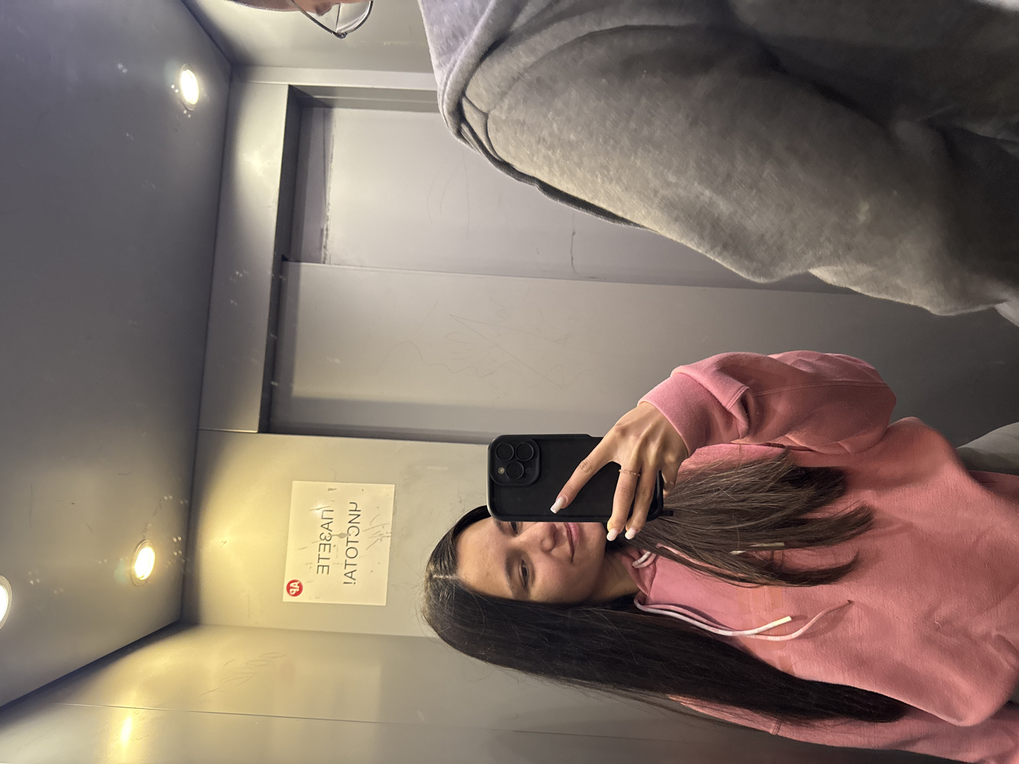 A selfie is taken in a vertically oriented image inside an elevator. A person with dark, long hair, wearing a pink hooded sweatshirt, holds a black smartphone. The camera lens is visible on the phone's back. The person appears to be looking at the phone screen, perhaps composing the photo.
A second person, only partially visible, wears a grey hoodie. The scene is an elevator cabin with metallic walls. A sign with lettering in Bulgarian, oriented vertically, is on the wall. The location is presumed to be in Varna, Bulgaria. The elevator has recessed lighting.
