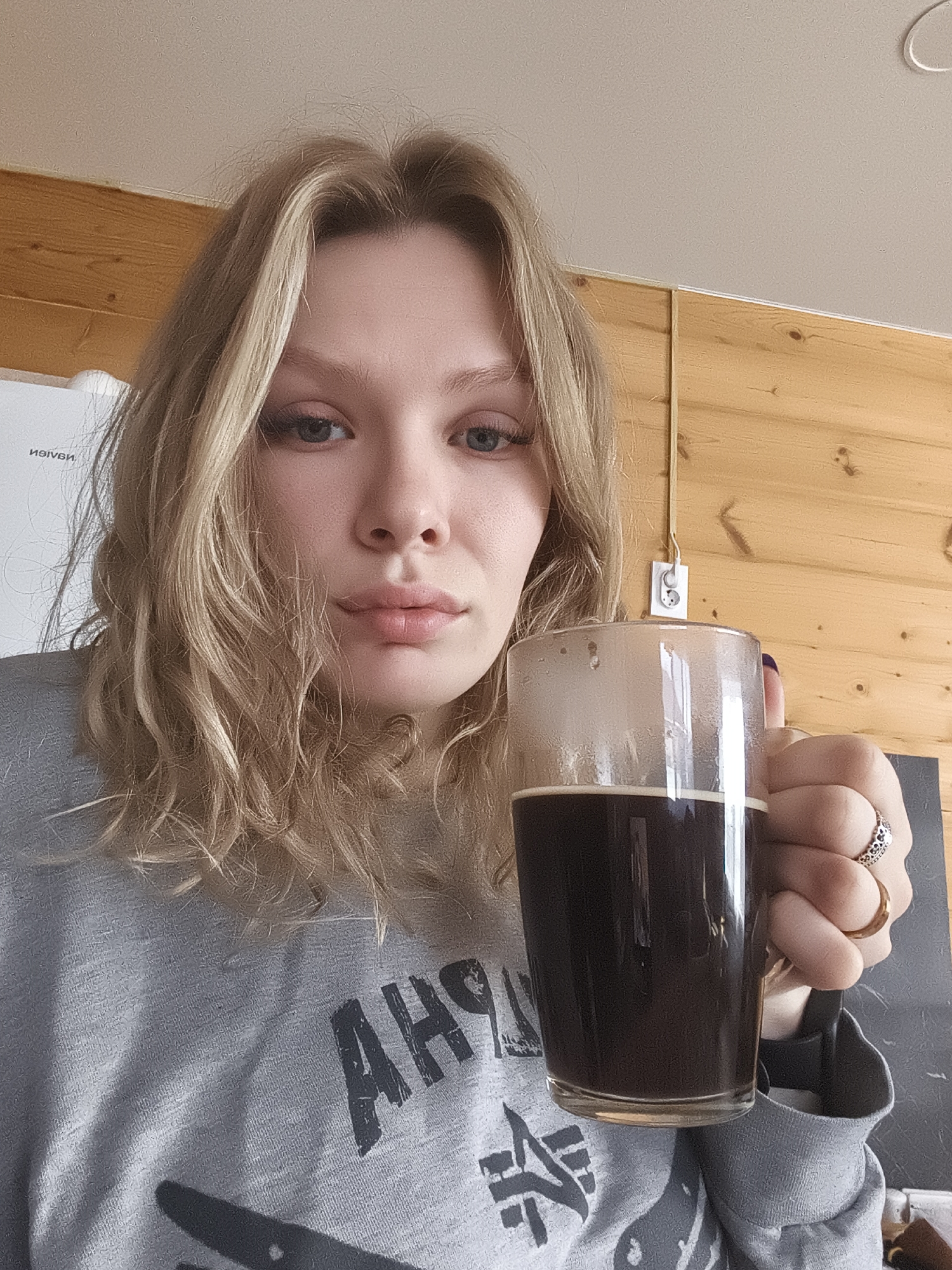 A blonde-haired woman is holding a clear glass mug filled with a dark liquid, possibly coffee or tea. She is wearing a grey long-sleeved shirt with a partially visible graphic design. She is wearing a ring on her right hand. Behind her, there is a wooden panel wall with an electrical outlet. A white board is visible to the left. The image does not provide enough information to determine whether it was taken in Русский Вожой, Russia.