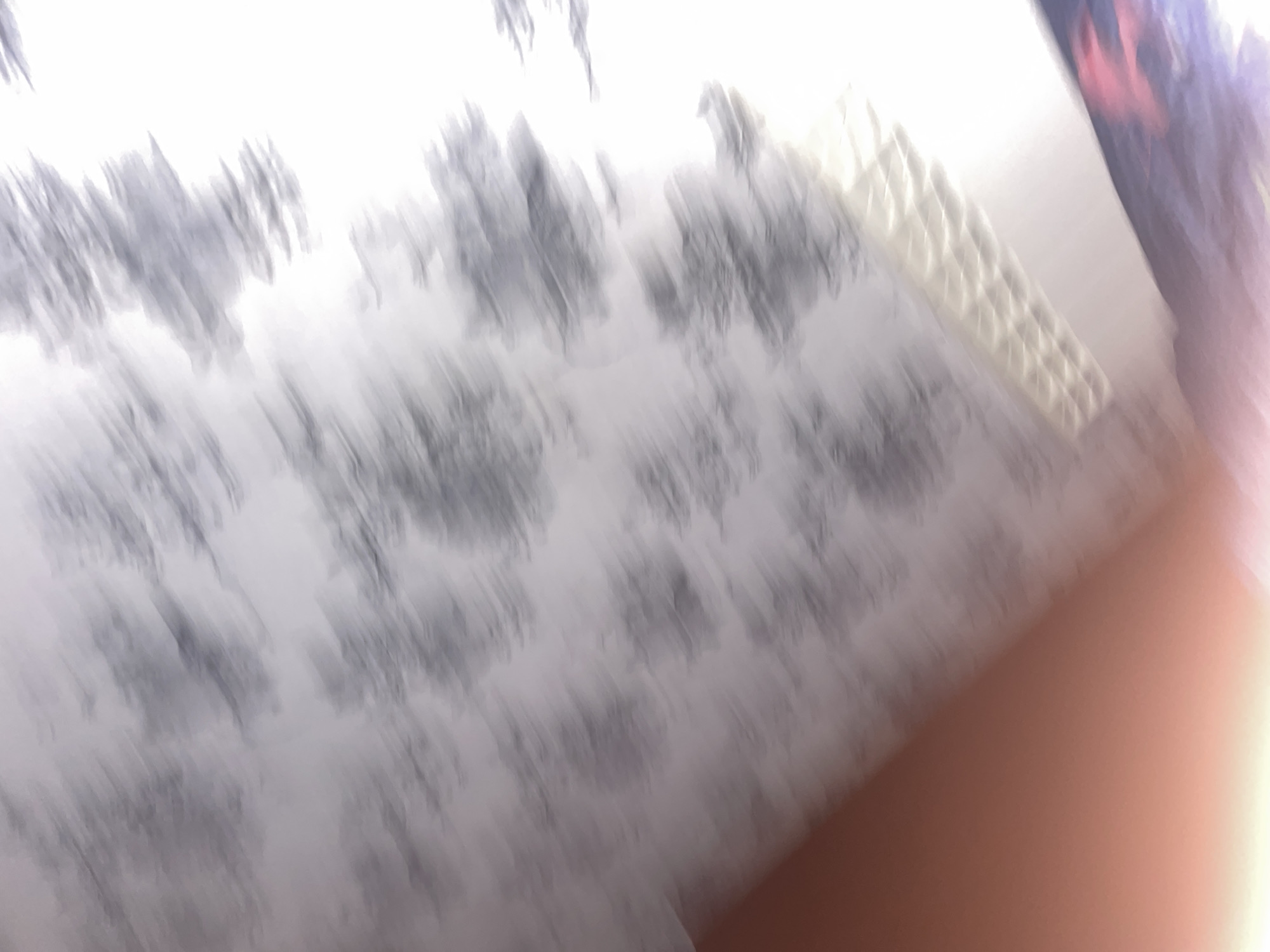 The image is a blurry, tilted photograph of what appears to be a ceiling with a patterned wallpaper. There is a strong orange cast on the lower left, possibly from a light source or lens flare. A portion of what looks like a light fixture or textured surface is visible in the lower right. The overall impression is of an indoor environment, but the blurriness and lack of distinct features make it impossible to identify specific objects or activities. The location within Атяшевское городское поселение, Russia, cannot be determined from the visual information provided.
