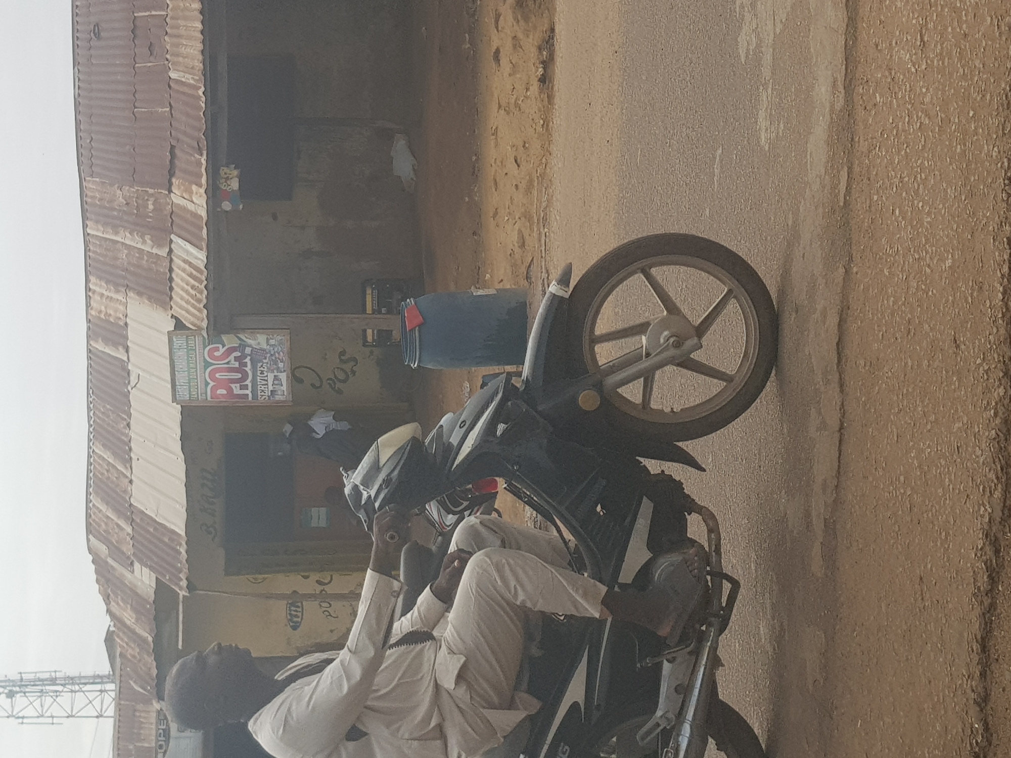 The image depicts a street scene in Kufena, Nigeria. A man dressed in white is sitting on a motorcycle parked adjacent to a building. The building has a corrugated iron roof and a sign reading "POS Service" is visible. A blue container sits next to the building. The street is unpaved.