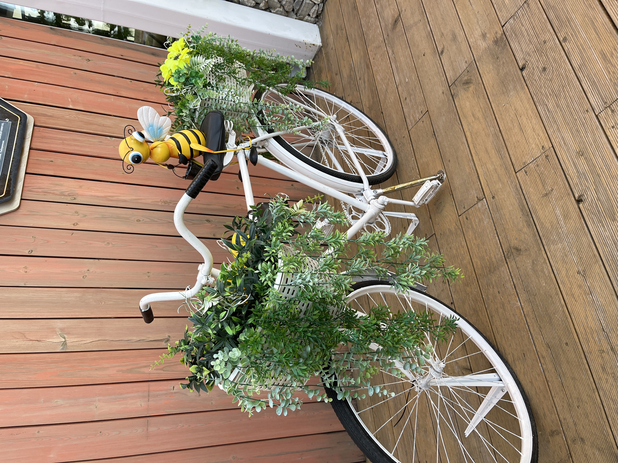 The image displays a decorative white bicycle adorned with artificial plants and a bee figurine, positioned on a wooden deck. A prominent yellow and black striped bee figurine, featuring white wings and black antennae, is attached to the bicycle's handlebars, facing left. The bicycle's front and rear baskets are filled with a lush arrangement of artificial green foliage and some yellow flowers, giving the impression of a vibrant, living display. The bicycle itself has a white frame and white-spoked wheels, contrasting with its black tires and gold-colored pedal components.
The bicycle rests on a light brown wooden deck, characterized by parallel planks showing some signs of wear. In the background, a white structure, likely a railing or part of a building, runs along the edge of the deck. Beyond this white structure, a section of stacked stones or rocks is visible. The scene is brightly lit by natural daylight, indicating clear weather. No people are present, and no activity is occurring; the image captures a static, decorative outdoor arrangement. A small, black sign with fragmented white text, possibly including Cyrillic characters, is partially visible on the left side of the frame, but the text is too indistinct to be read. This charming display is situated in an outdoor area in Chaikovsky, Russia.
